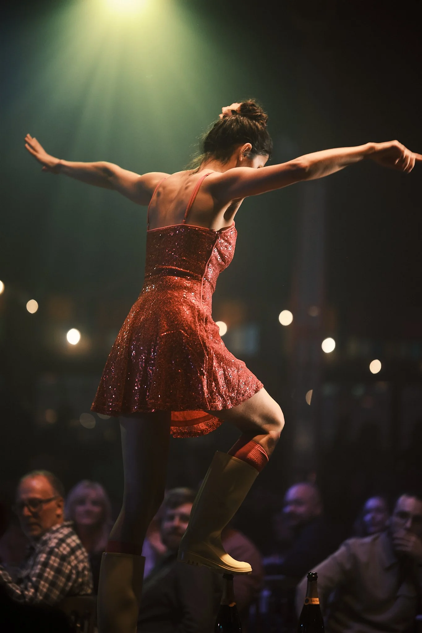 A woman in a sparkly red dress and yellow rain boots balancing on a beer bottle, with her arms outstretched, during a performance in front of an audience at night.