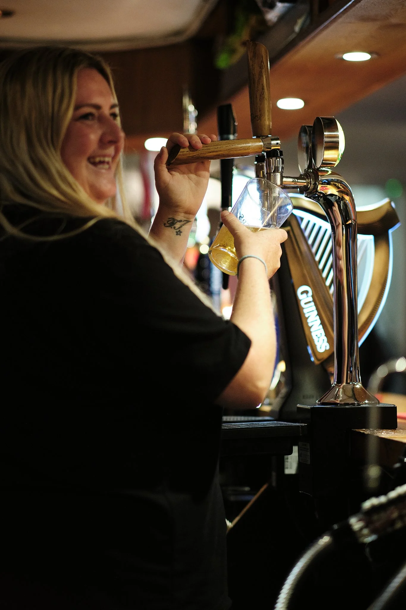 A woman with long blonde hair smiling while pouring a draft beer from a tap at a bar.