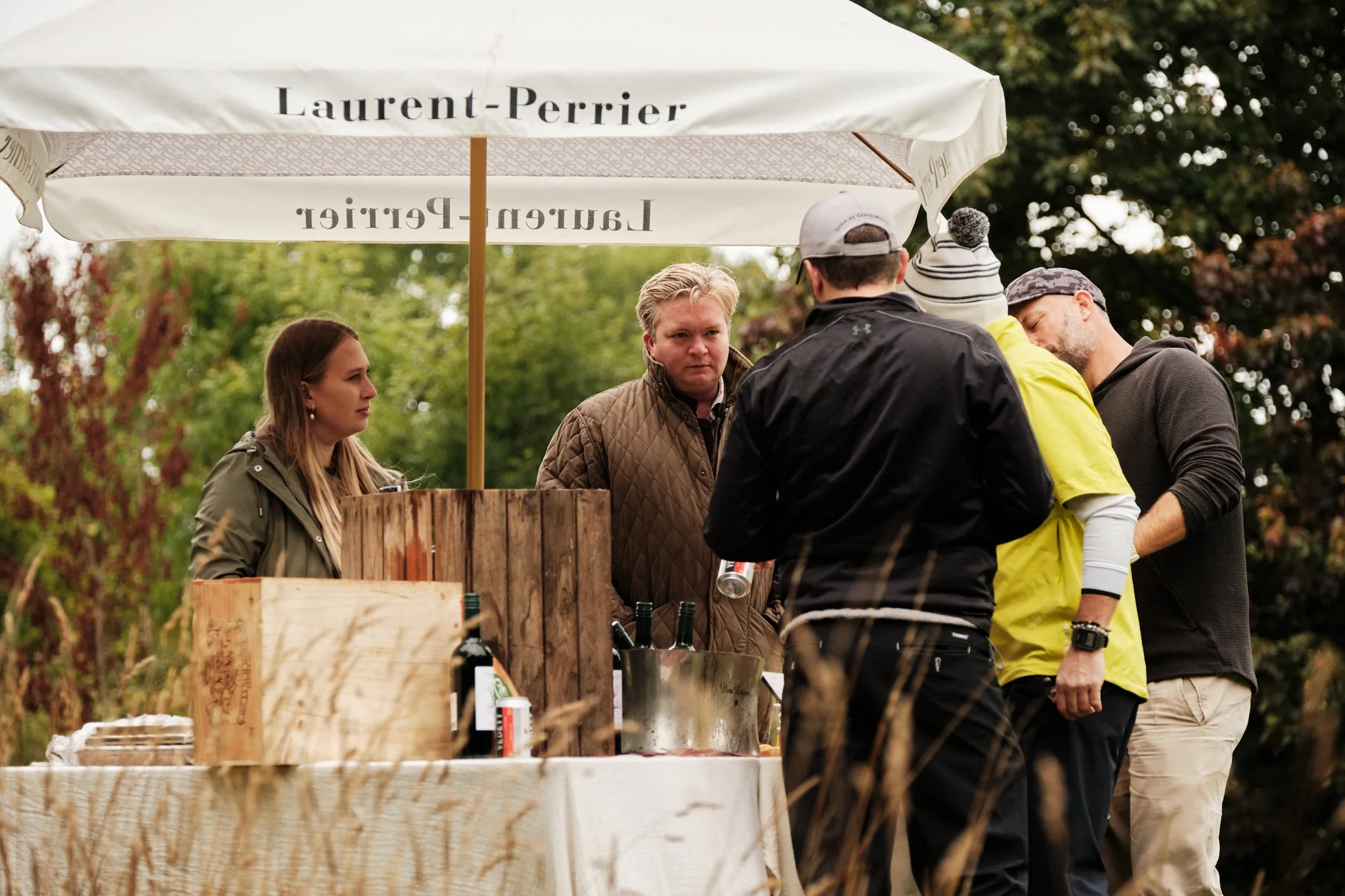 Group of five people gathered under Laurent-Perrier umbrella at an outdoor event, engaging in conversation near a table with wine bottles and food items, surrounded by trees.
