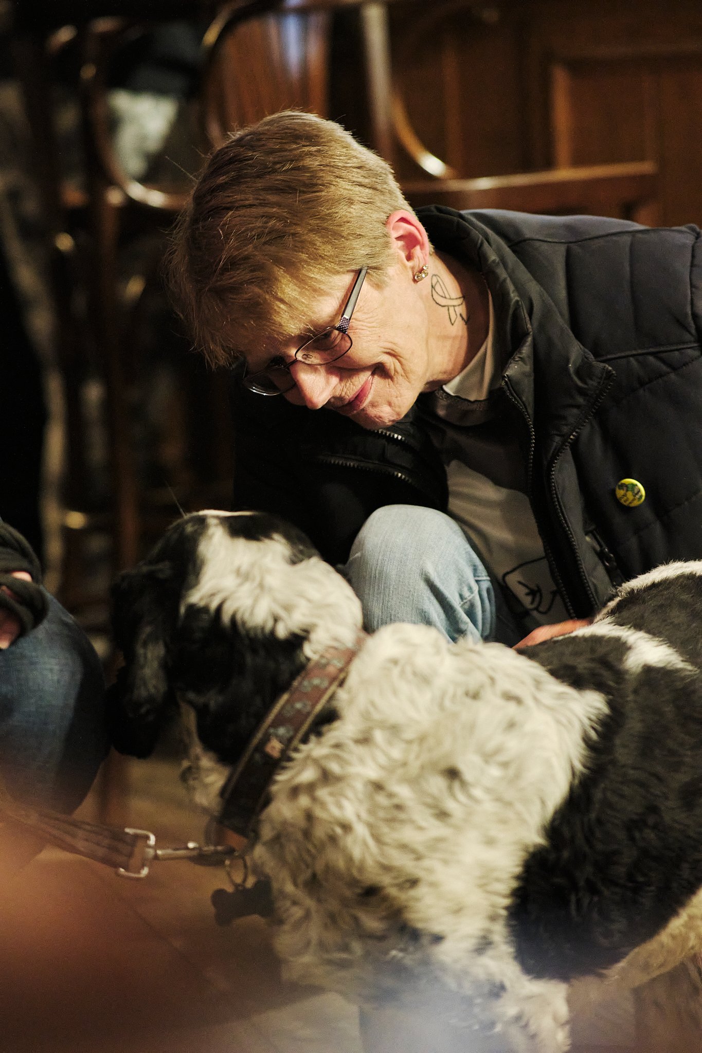 Older woman with short hair, glasses, and tattoos on neck and ear, smiling and leaning towards two black and white puppies on the floor in a warm, cozy indoor setting.