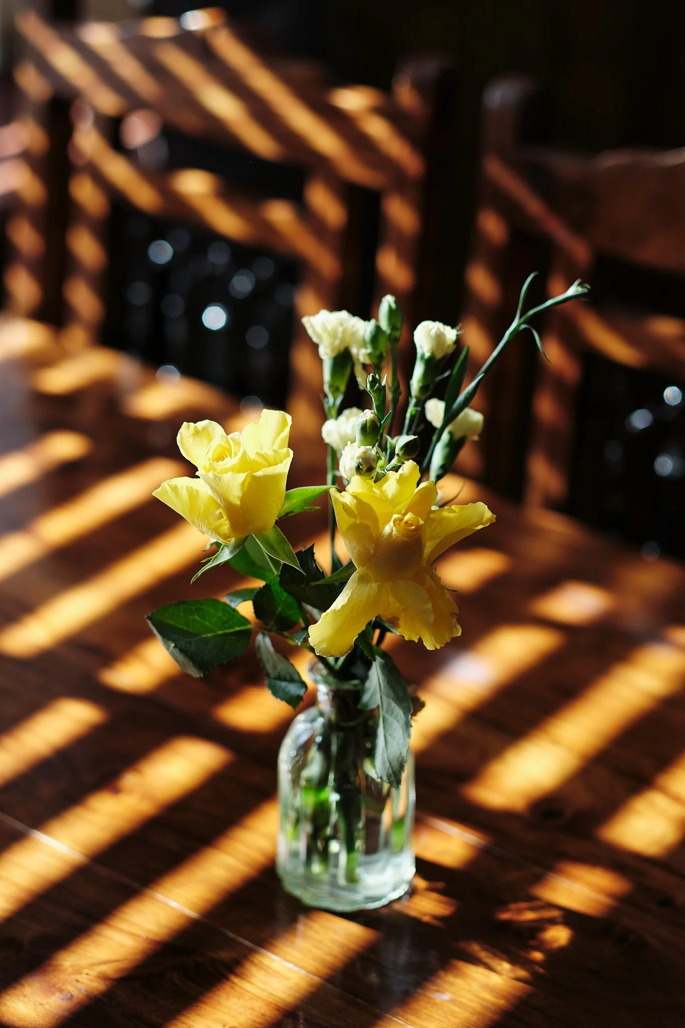 A small glass vase with yellow and white flowers sitting on a wooden table with sunlight casting striped shadows.