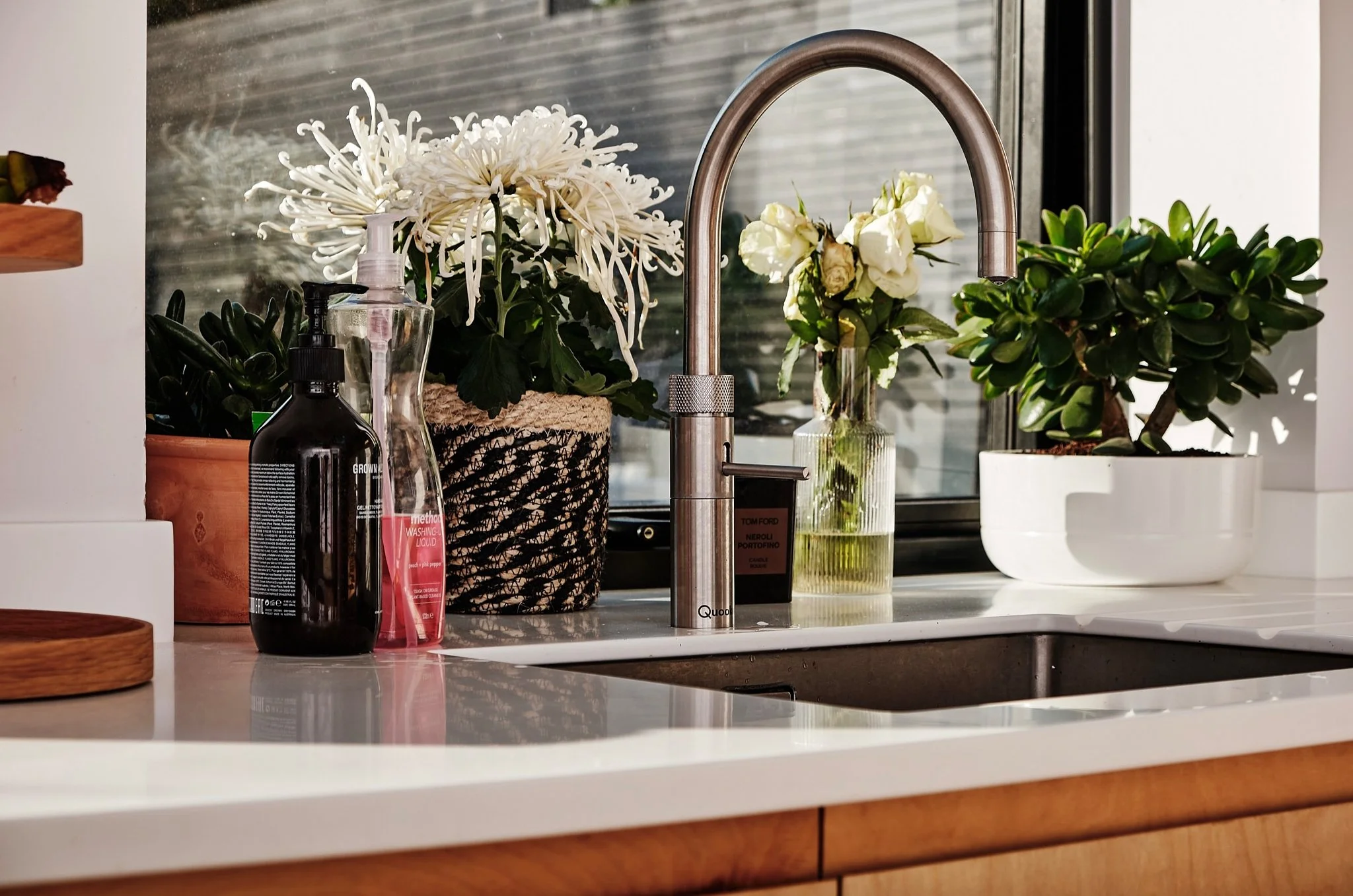 Kitchen sink with potted plants and flowers, soap dispenser, and liquid hand soap, set against a window with blinds.