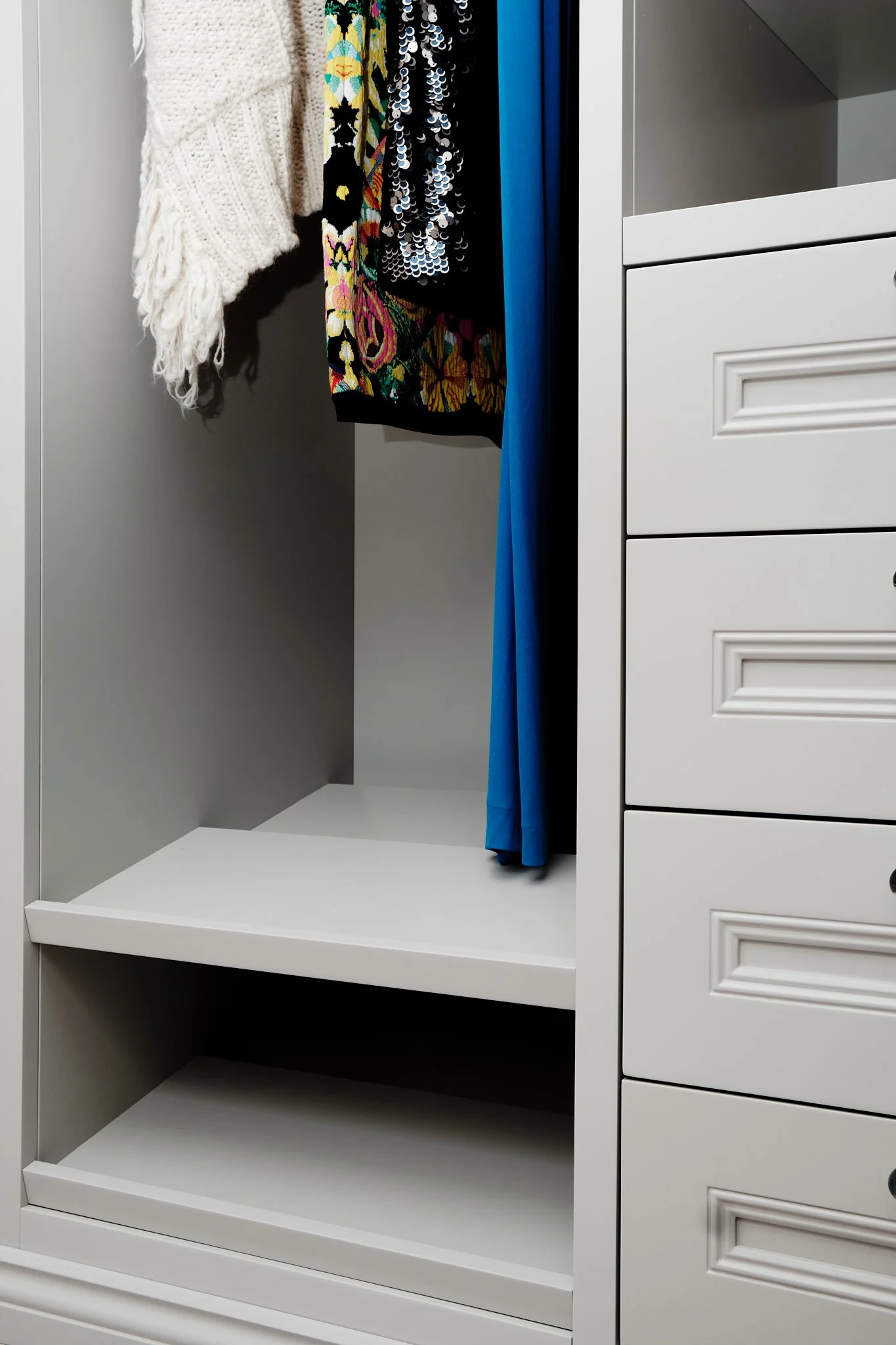 Empty closet with hanging clothes including a white knitted sweater, a multicolored floral dress, a black sequin dress, and a blue garment, with shelves and drawers on the right side.