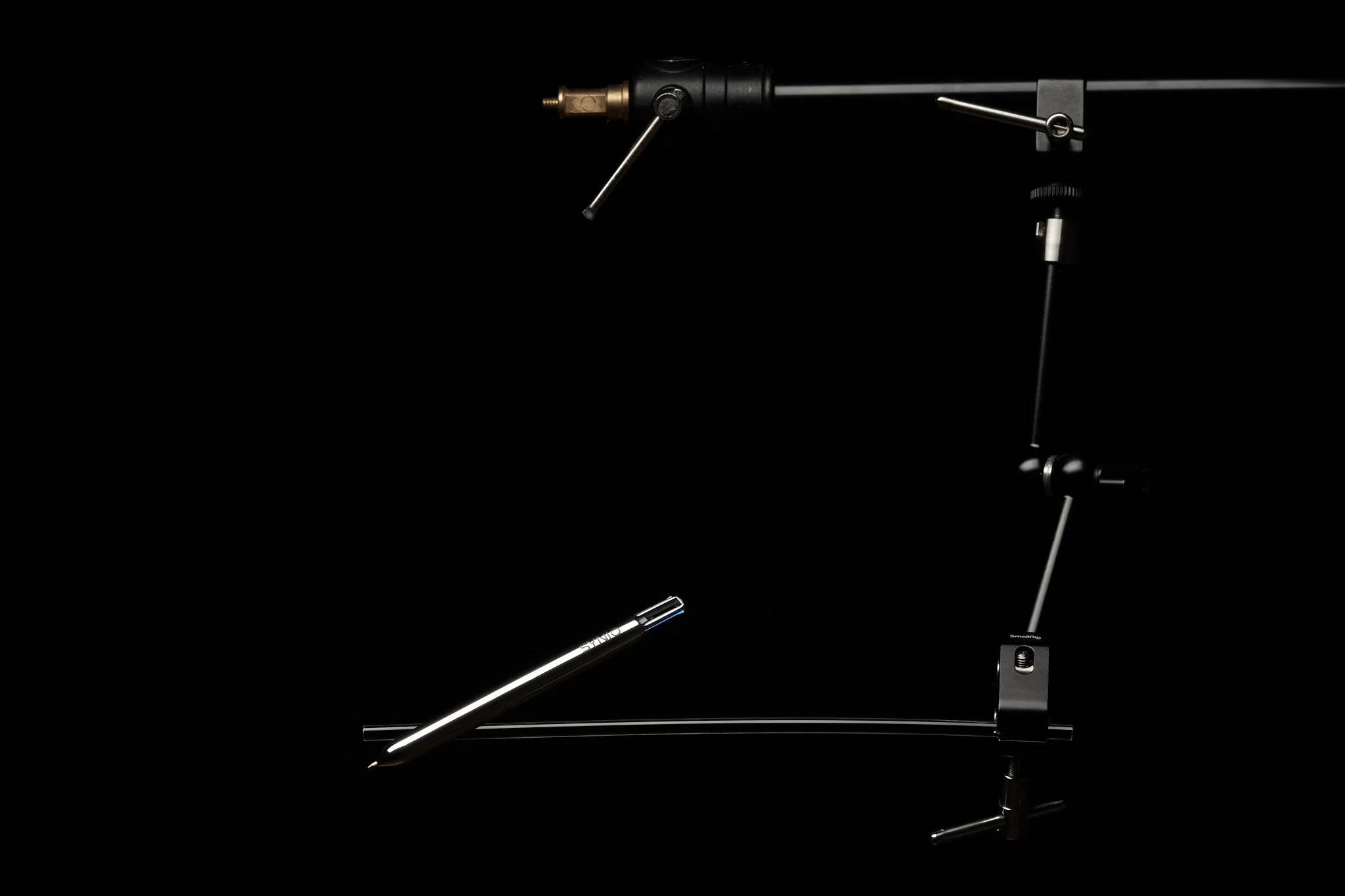 A turntable tonearm with a pen positioned underneath, set against a black background.