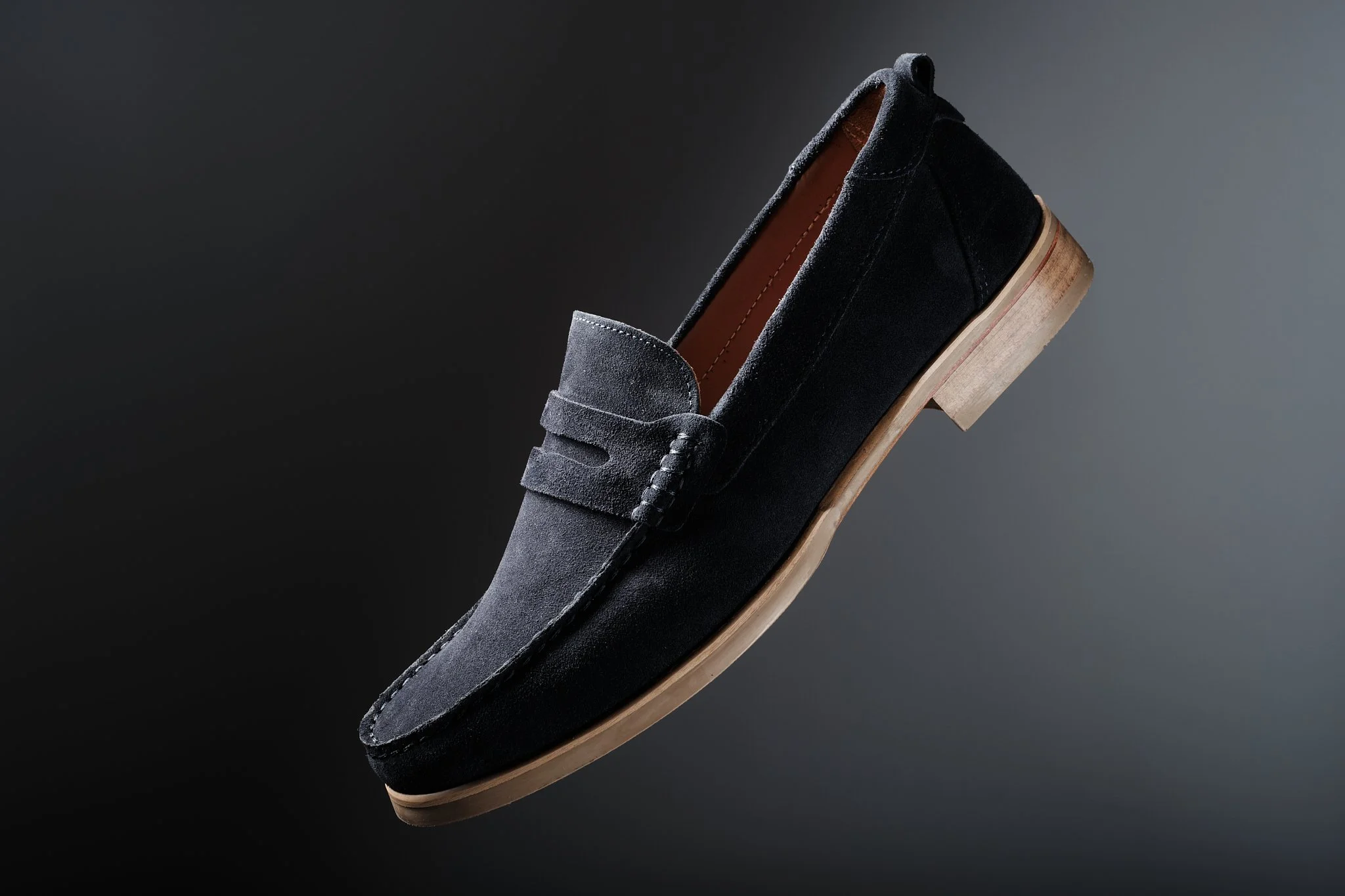 A black suede men's loafer shoe with a wooden sole and slight heel, isolated on a dark gradient background.
