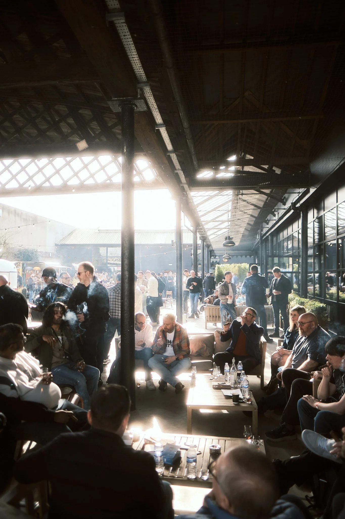 People gathered and socializing at an outdoor patio area with sunlight streaming through the roof, some seated and some standing, with tables, drinks, and a casual atmosphere.