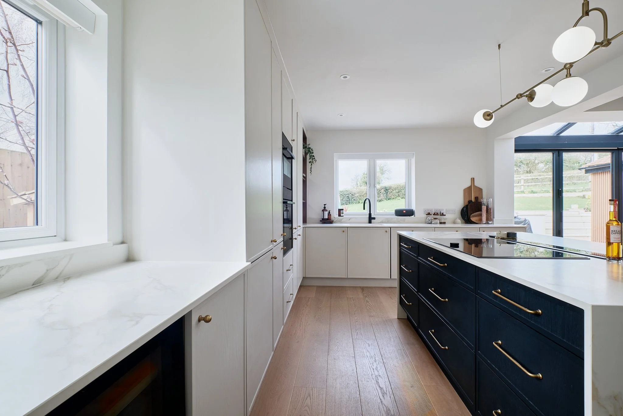 Modern kitchen with white cabinets, a black island with gold handles, wooden flooring, and large windows.