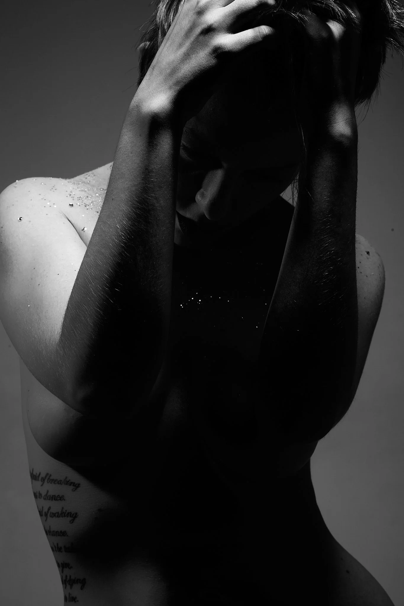 A black-and-white photograph of a woman with wet skin, covering her face with her hands, with water droplets visible on her shoulder and arm, and a tattoo on her left side.