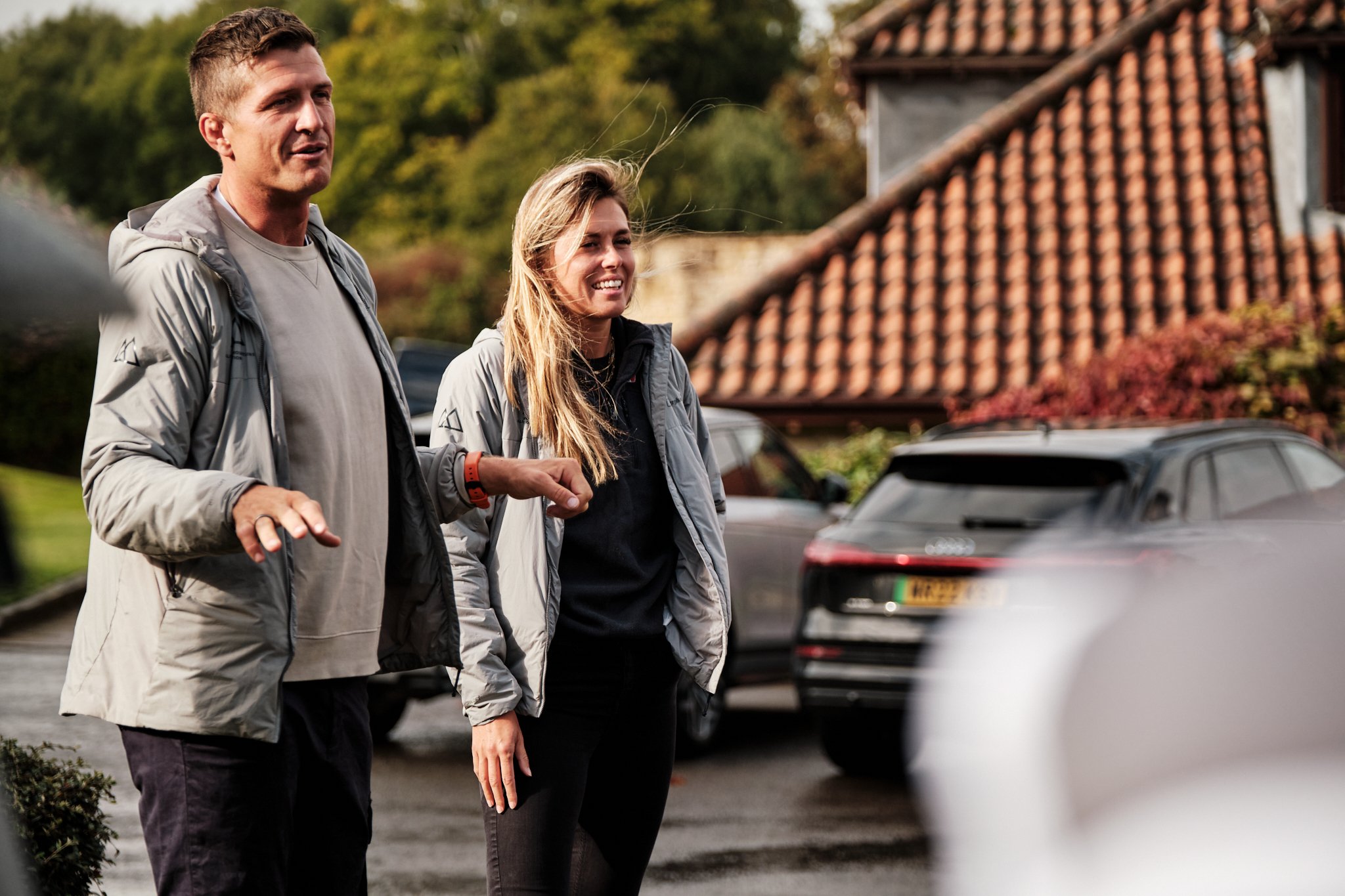 Two people standing outdoors in front of cars, engaged in conversation, with a house and trees in the background.