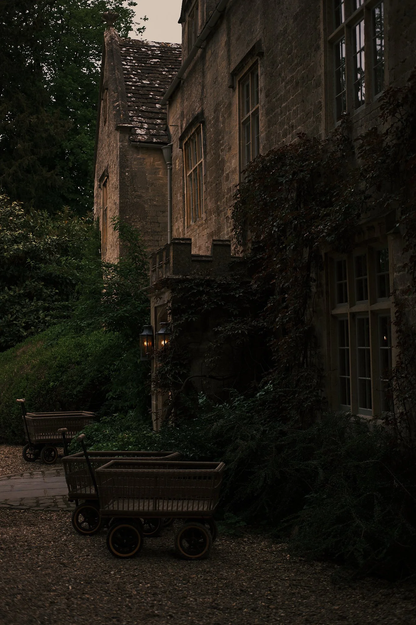 Dark, old stone building with multiple windows, surrounded by greenery and bushes, with two empty shopping carts on a gravel path in the foreground. Hotel interior photography at THE PIG-in the Cotswolds