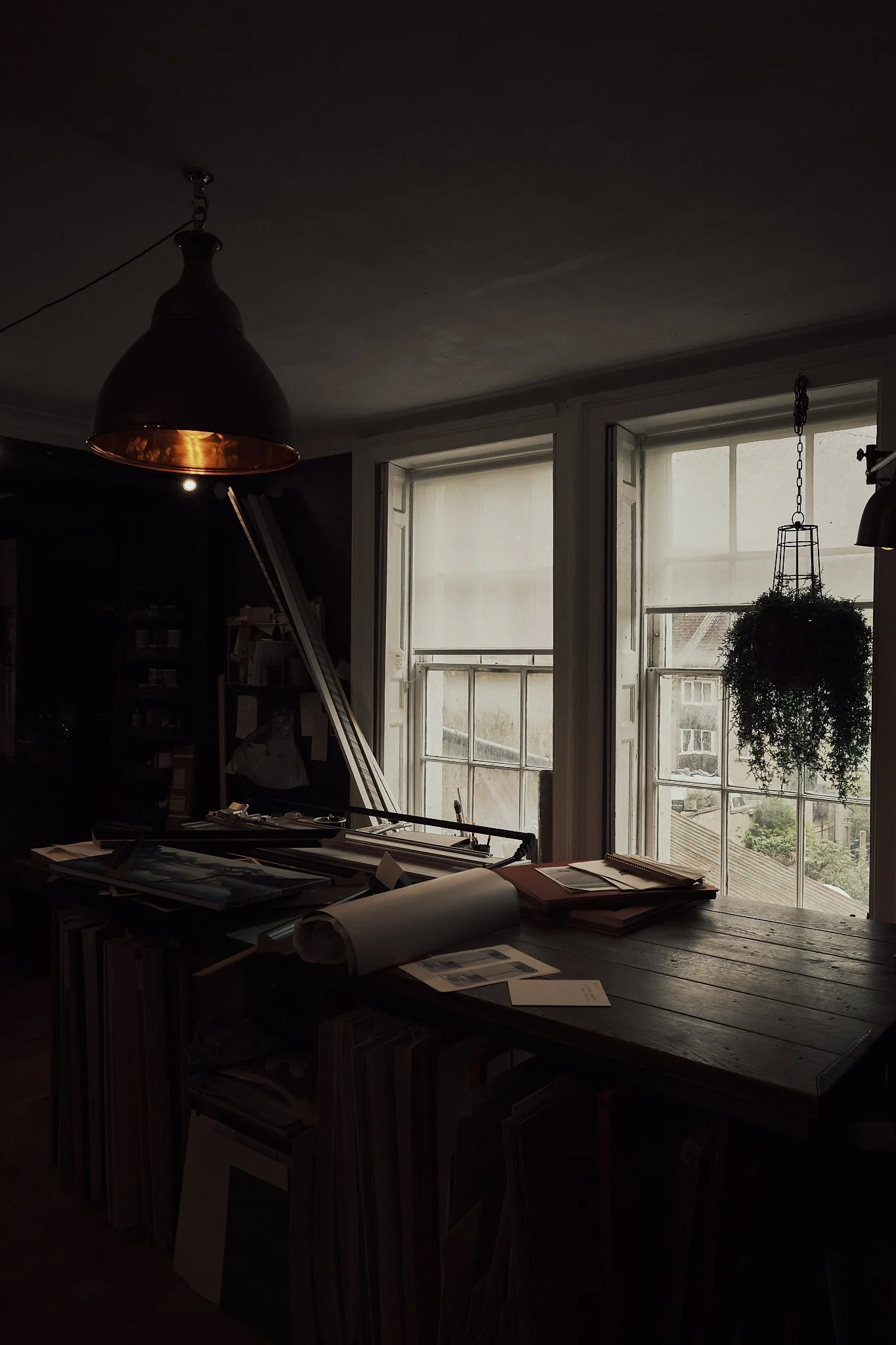 A dimly lit room with large windows letting in natural light, a wooden table cluttered with papers and documents, a hanging potted plant, and a black pendant lamp hanging from the ceiling.