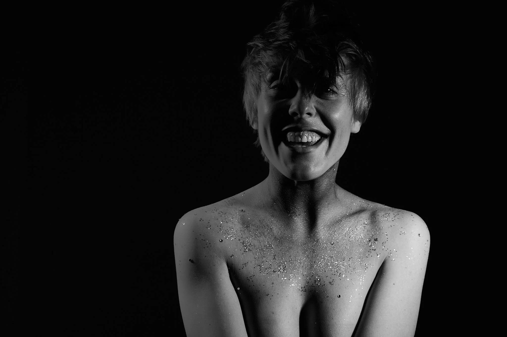 Black and white photo of a smiling person with messy hair, covered in glitter on their shoulders and chest, against a dark background.
