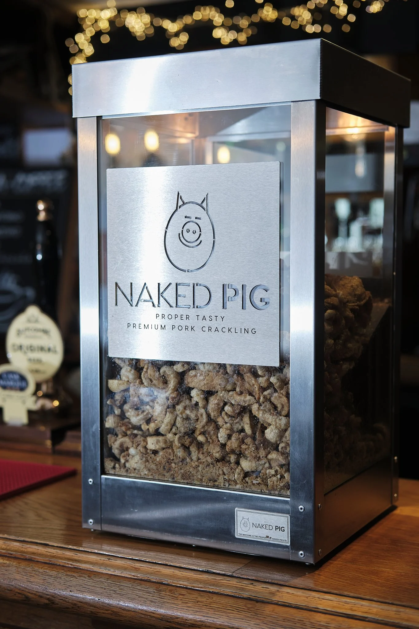 A metal display case filled with pork cracklings, labeled 'Naked Pig, Proper Tasty Premium Pork Crackling' with a pig face illustration on the front.