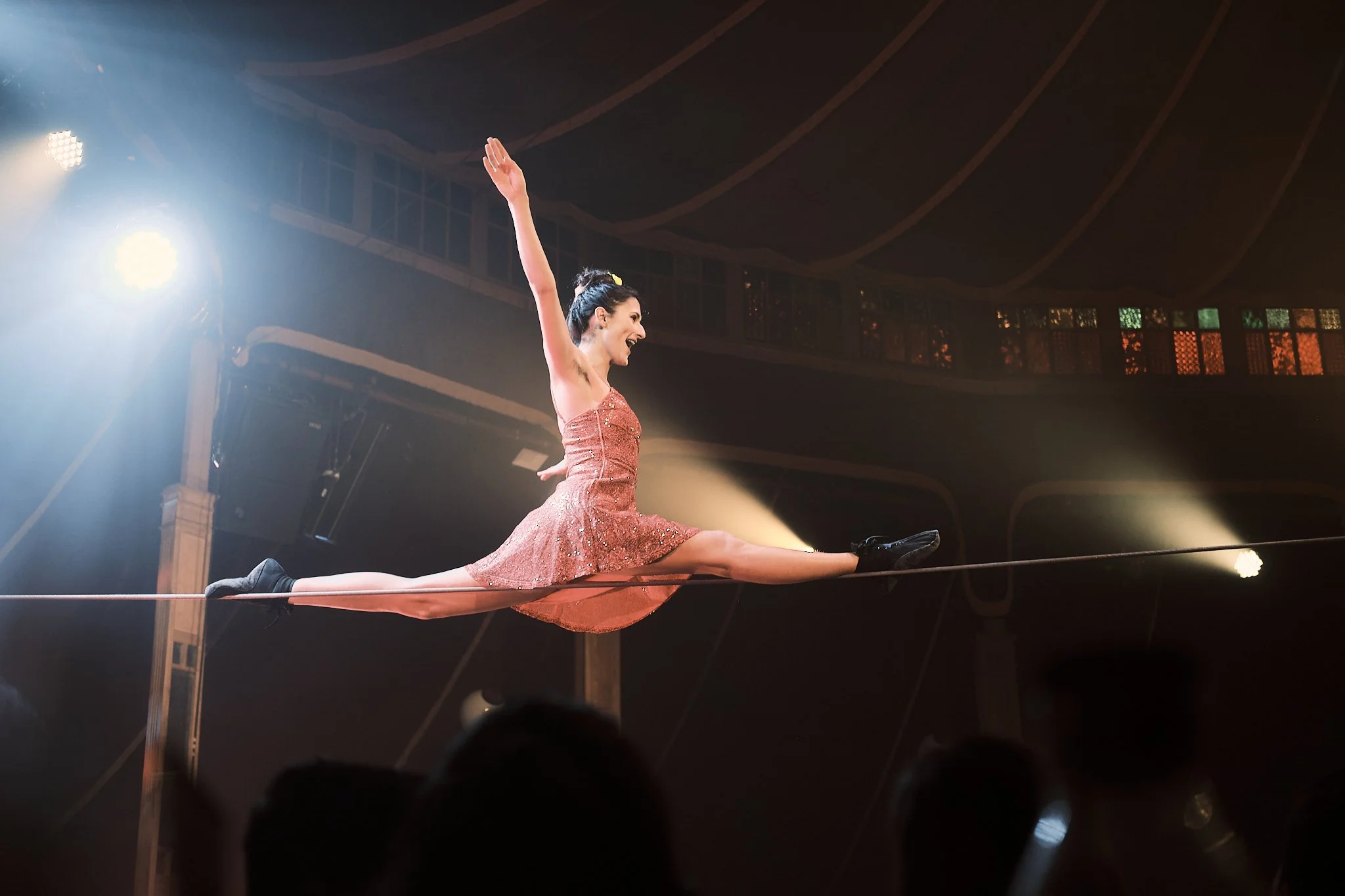 A female aerialist performing a split on a tightrope in a circus tent, dressed in a sparkly pink costume, with audience visible in silhouette at the bottom.
