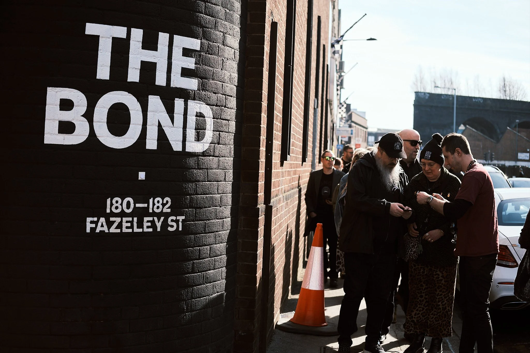 People standing in line outside a building with a large sign that reads 'The Bond', with additional text indicating the address '180-182 Fazeley St'.
