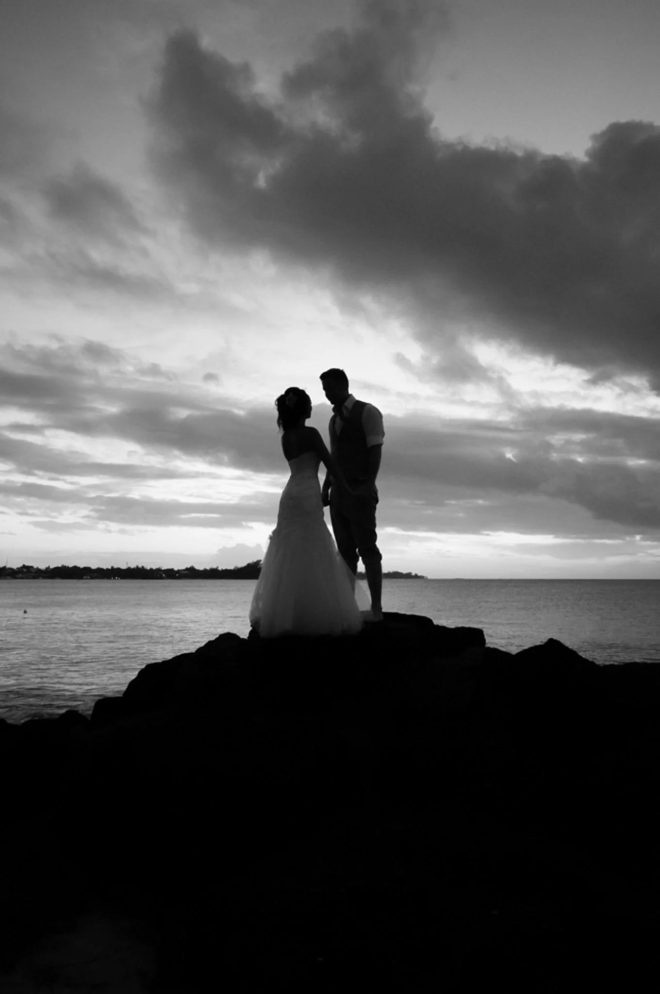 Silhouette of a couple in wedding attire standing on rocks by the water during sunset or dusk, with dark clouds in the sky.