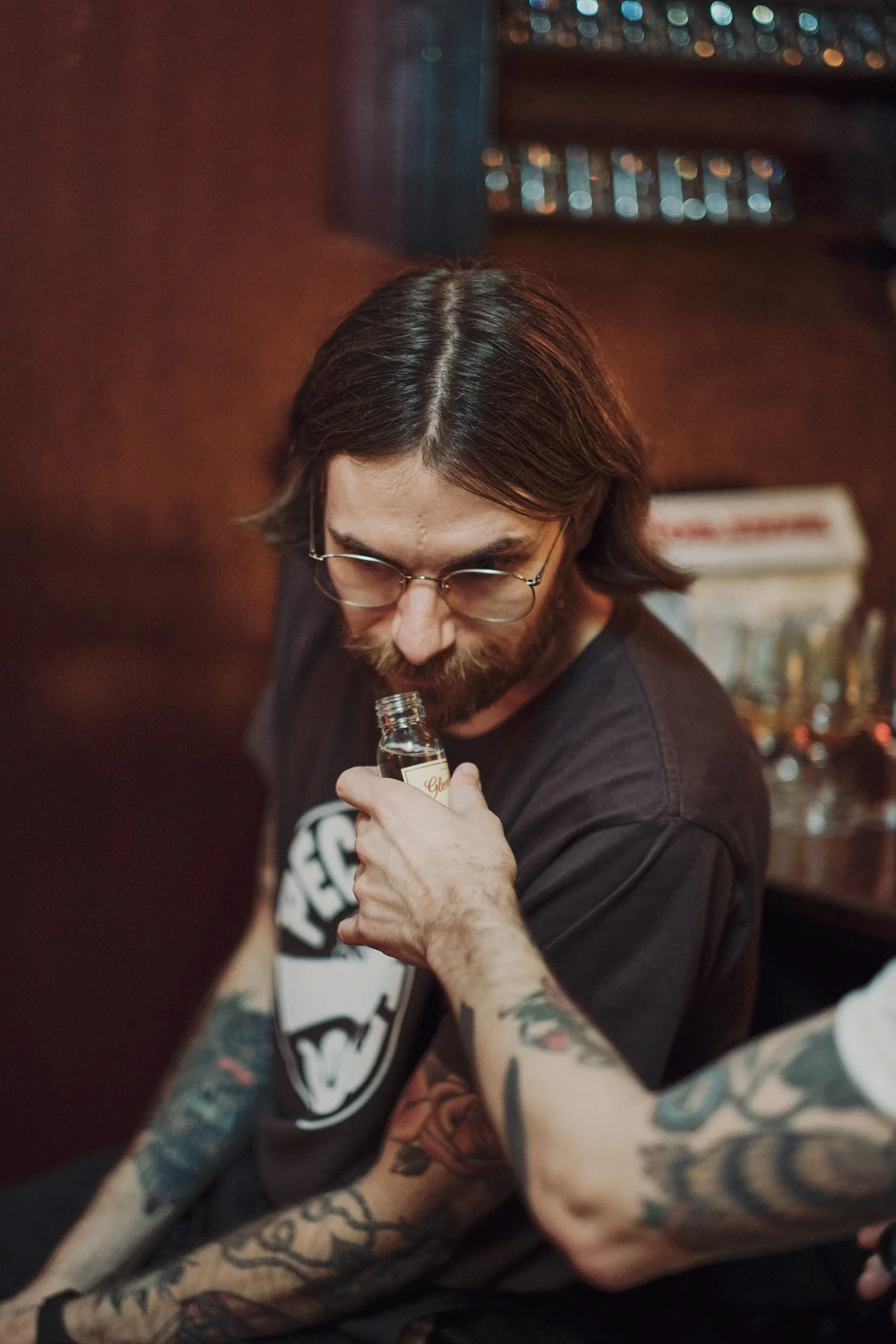 A man with long hair, glasses, and tattoos on his arms, holding a small bottle and about to take a sniff, sitting in a dimly lit room with a red wall and a window in the background.