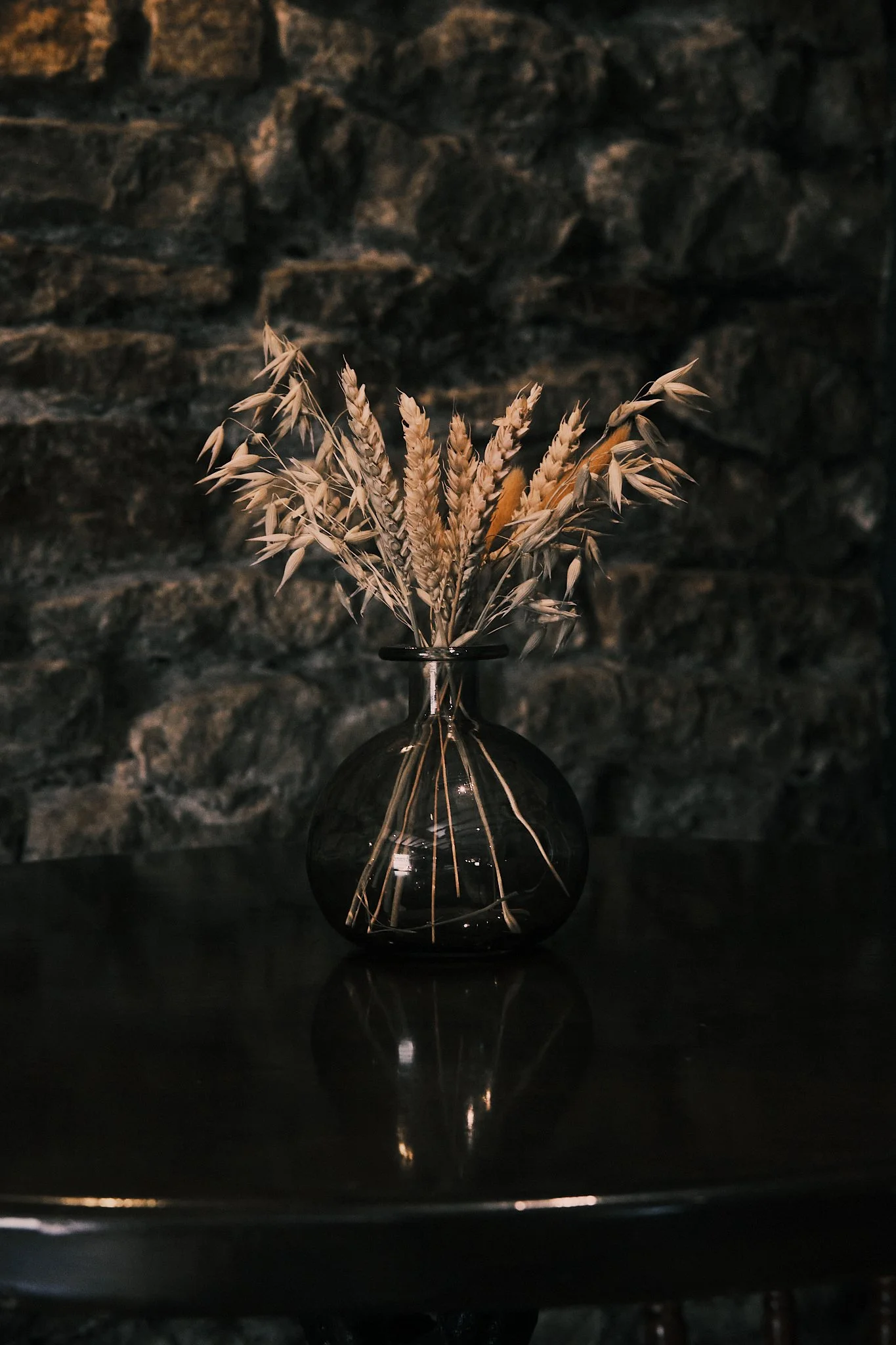 A black glass vase with dried wheat and grass stems, on a dark reflective surface against a textured stone wall background.