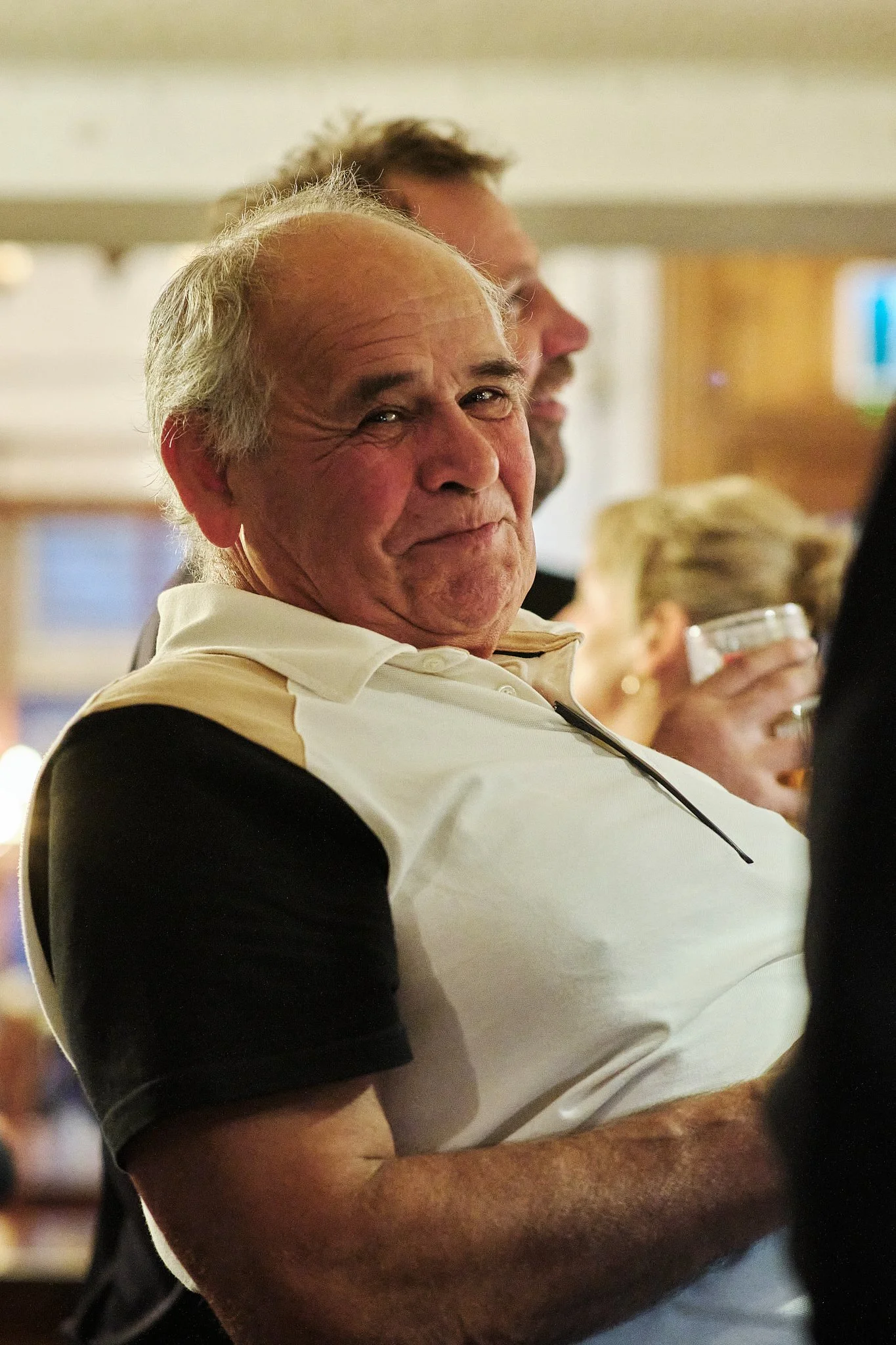 An older man with gray hair, wearing a white and black polo shirt, smiling and looking at the camera. He is at a social gathering with people in the background.