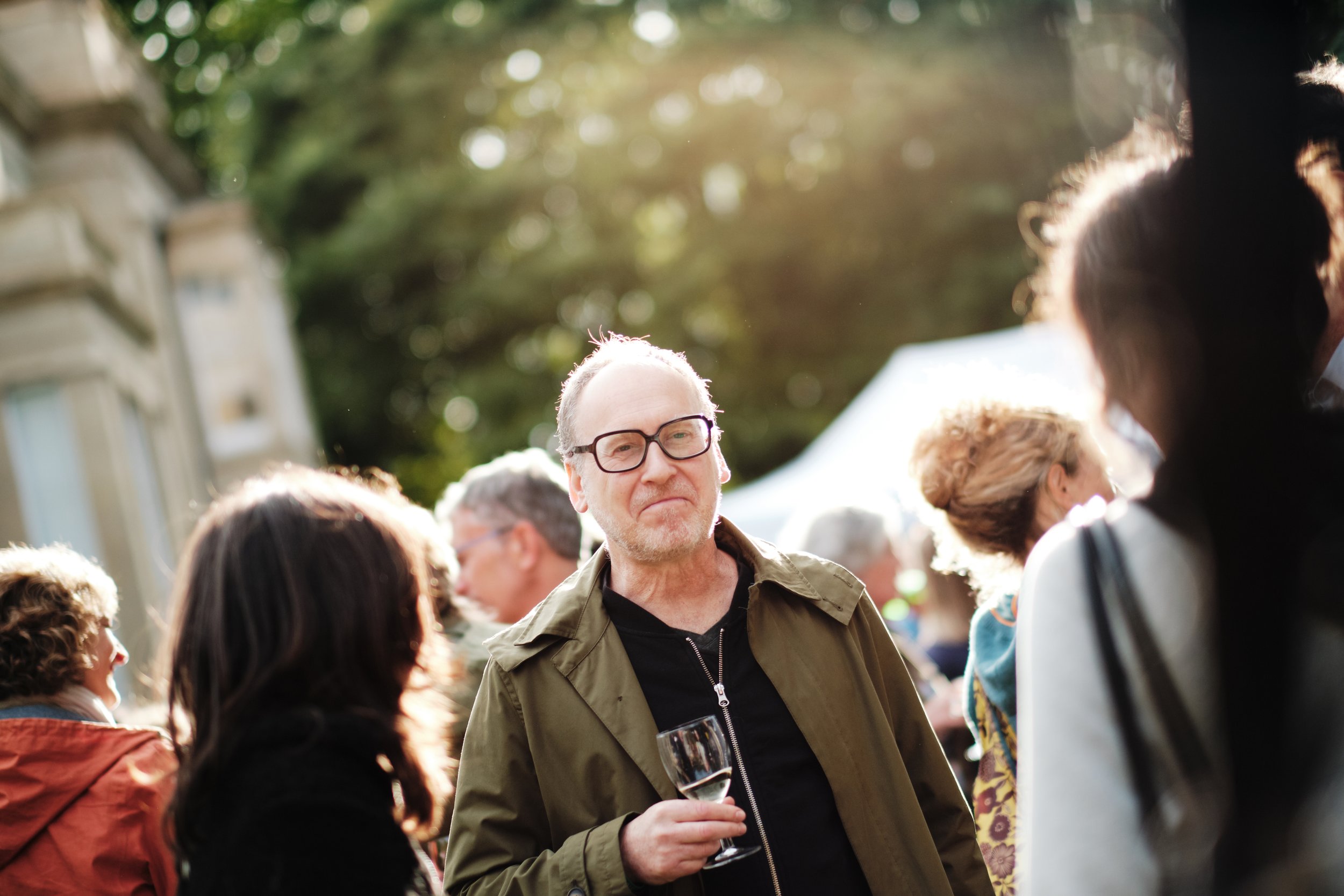 A man wearing glasses holding a wine glass at an outdoor event with other people around.