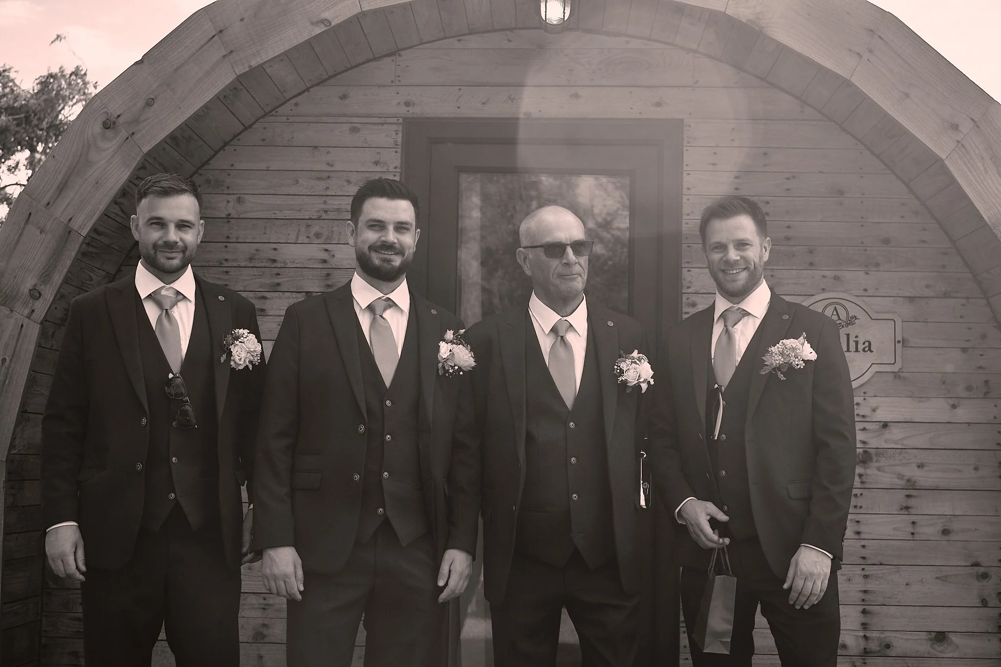 Four men in suits standing under a wooden archway in front of a wooden building. One man is older with sunglasses, and the others are younger, all smiling.