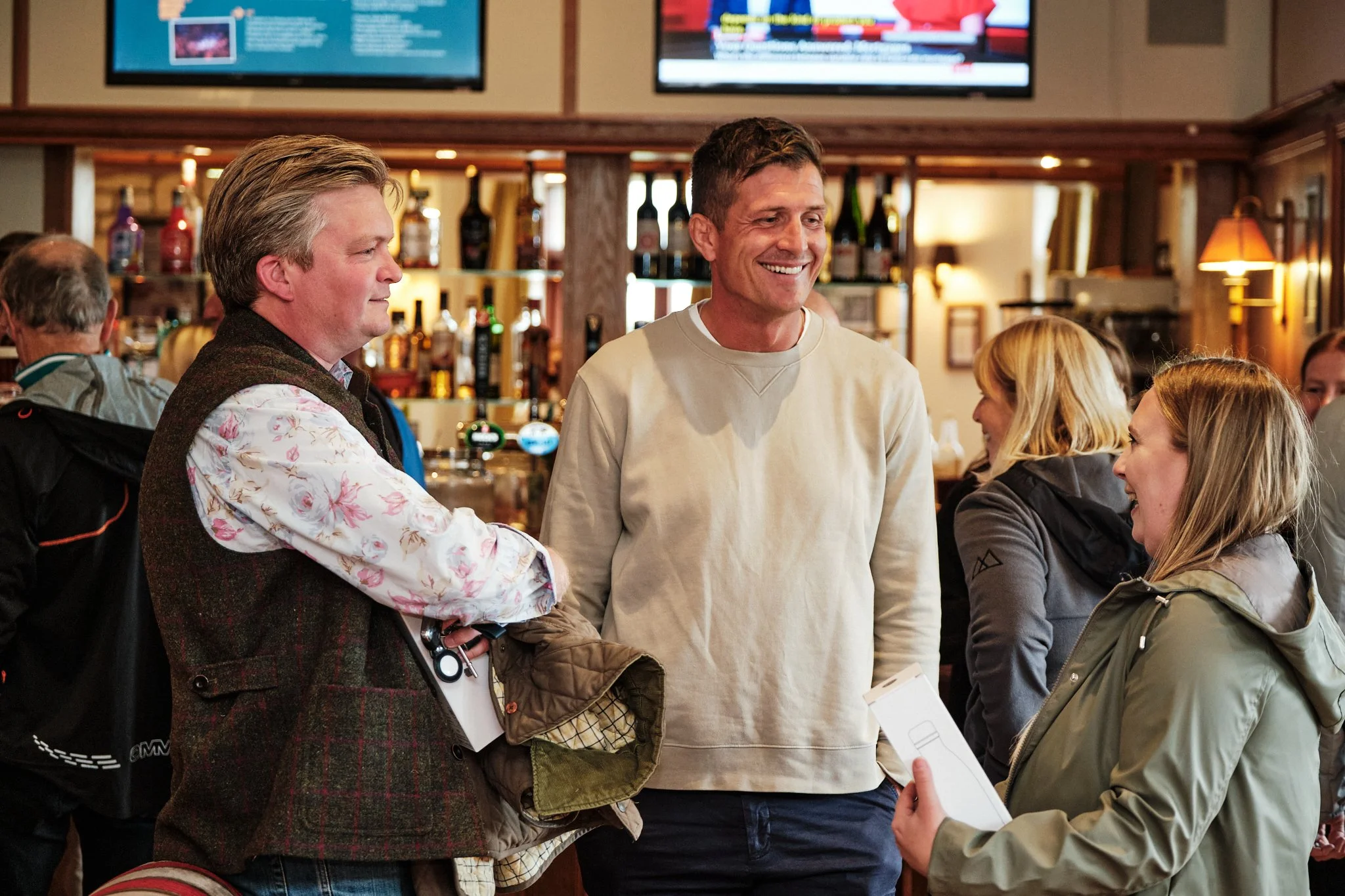 Three people engaged in conversation at a bar or pub, with others in the background and televisions on the wall.