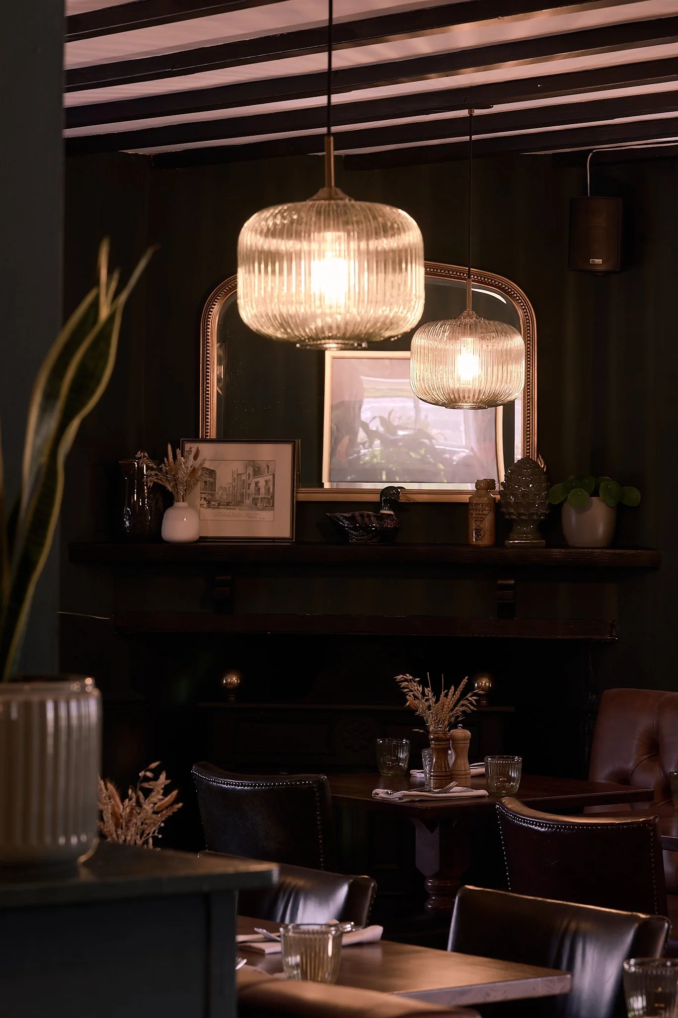 Interior of a dimly lit restaurant or cafe with hanging glass pendant lights, a mirror, framed artwork, a shelf with decorative items and plants, and tables set with glassware and napkins.