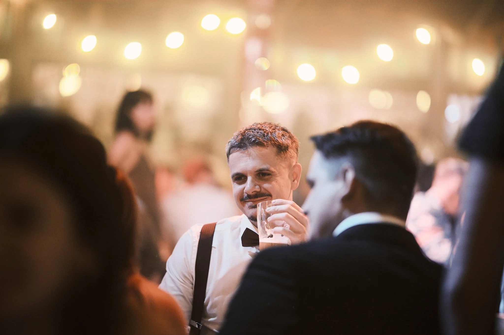 A man with a mustache and short hair, dressed in formal attire with a black bow tie and suspenders, is sitting at a social gathering or party, holding a glass of beer. The background is blurred with warm lighting and other guests.