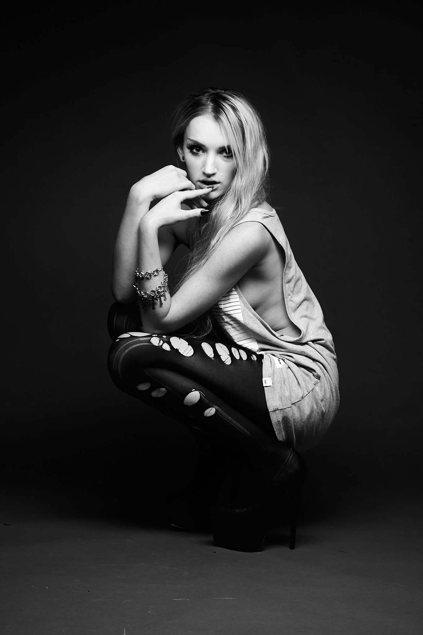 A black-and-white photo of a woman squatting against a dark background. She has long, wavy hair and is wearing a sleeveless top, patterned tights, and high-heeled boots. She has bracelets on one arm and is resting her chin on her hand.