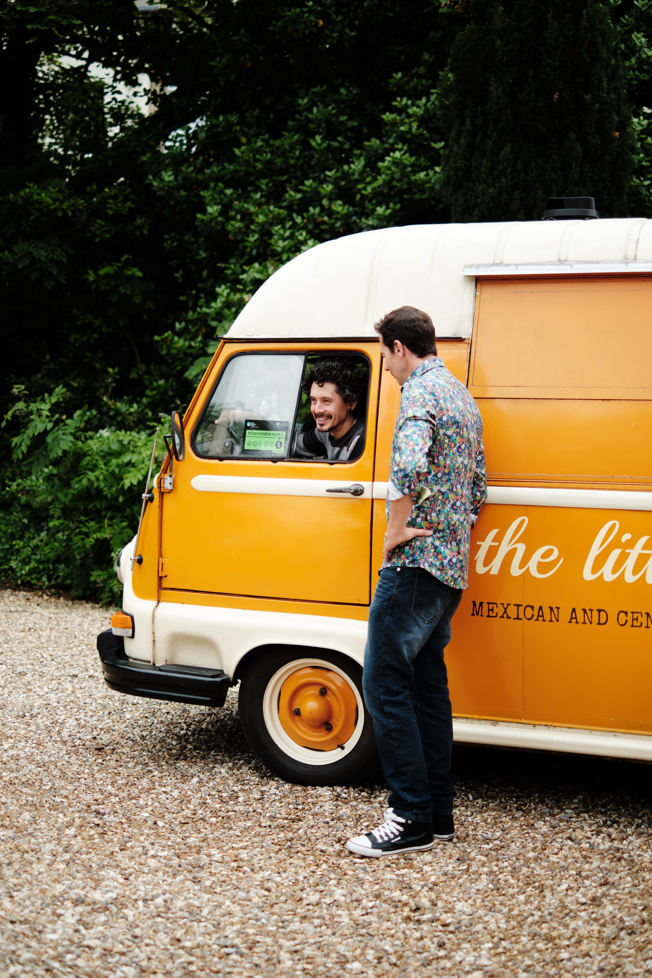 Two men conversing beside a vintage yellow food truck on a gravel surface, with green trees in the background.