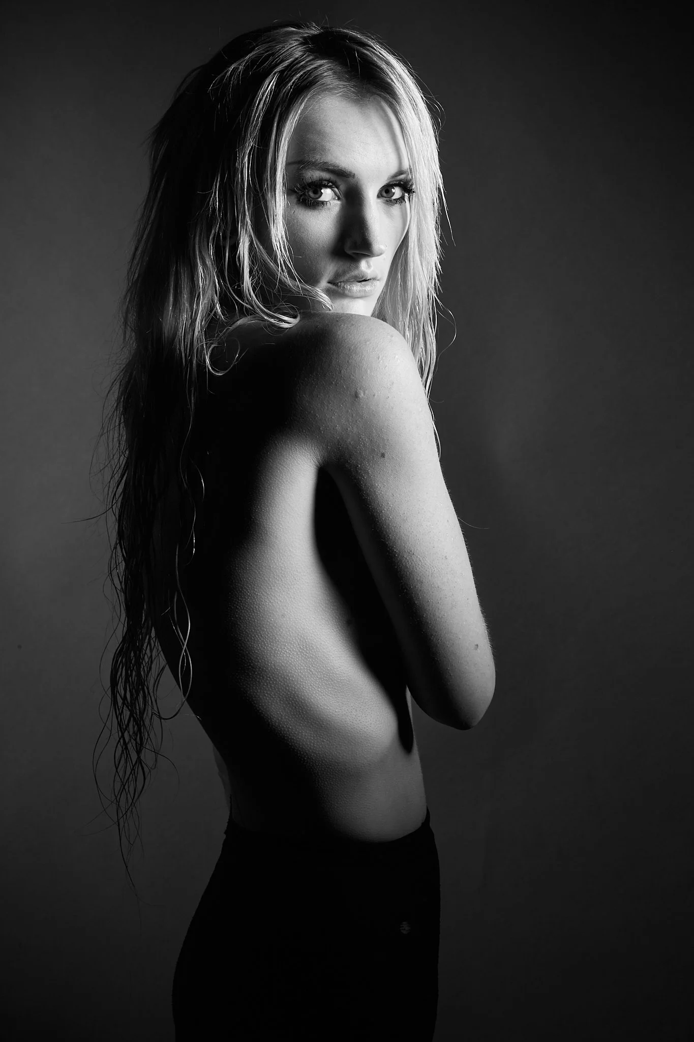 Black and white portrait of a young woman with wet hair, looking over her shoulder with a serious expression, topless, with her arms crossed over her chest.