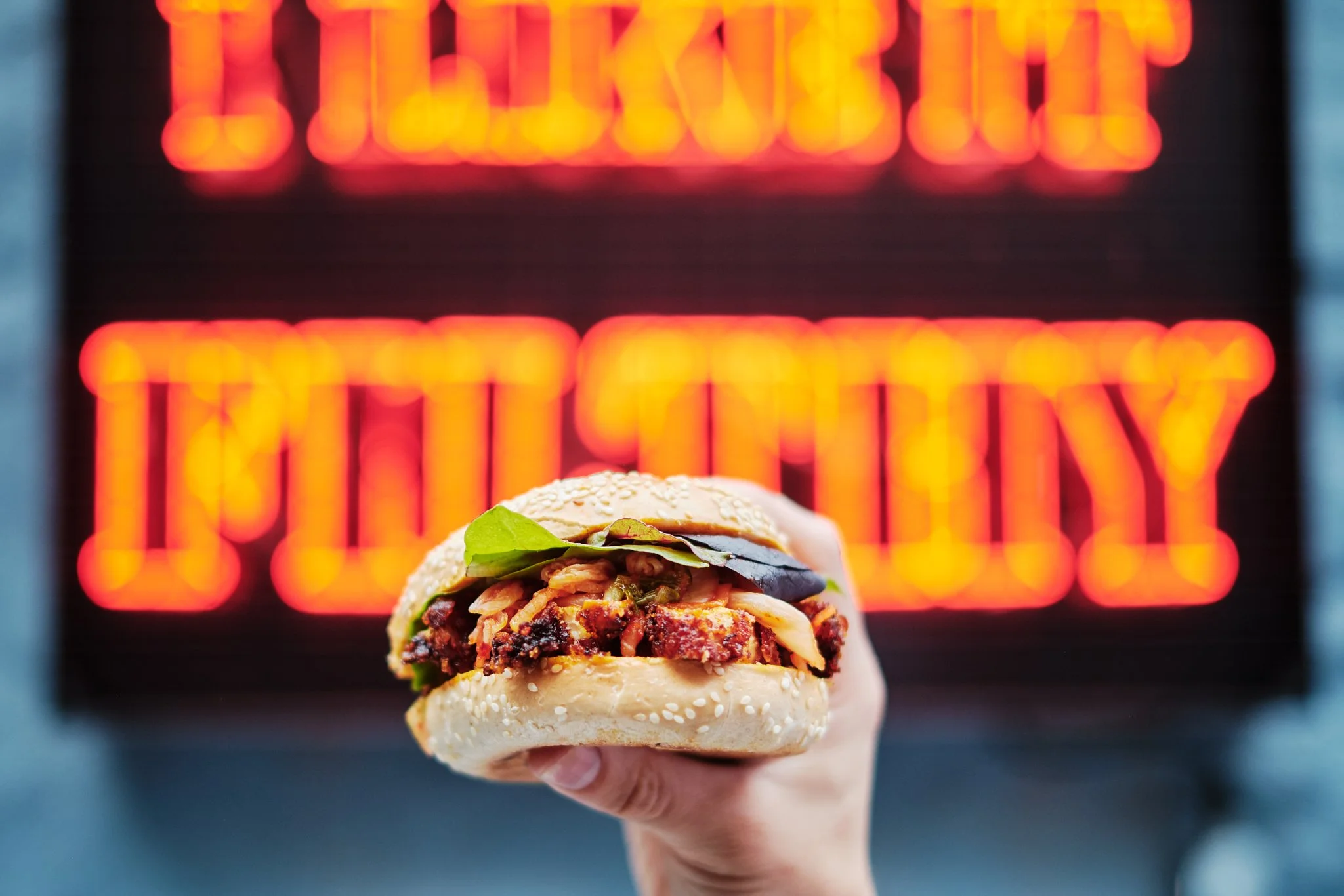 A hand holding a sandwich with shredded meat, lettuce, and sesame seed bun in front of a blurred neon sign that reads 'Lettuce Celebrate Tuesday'.