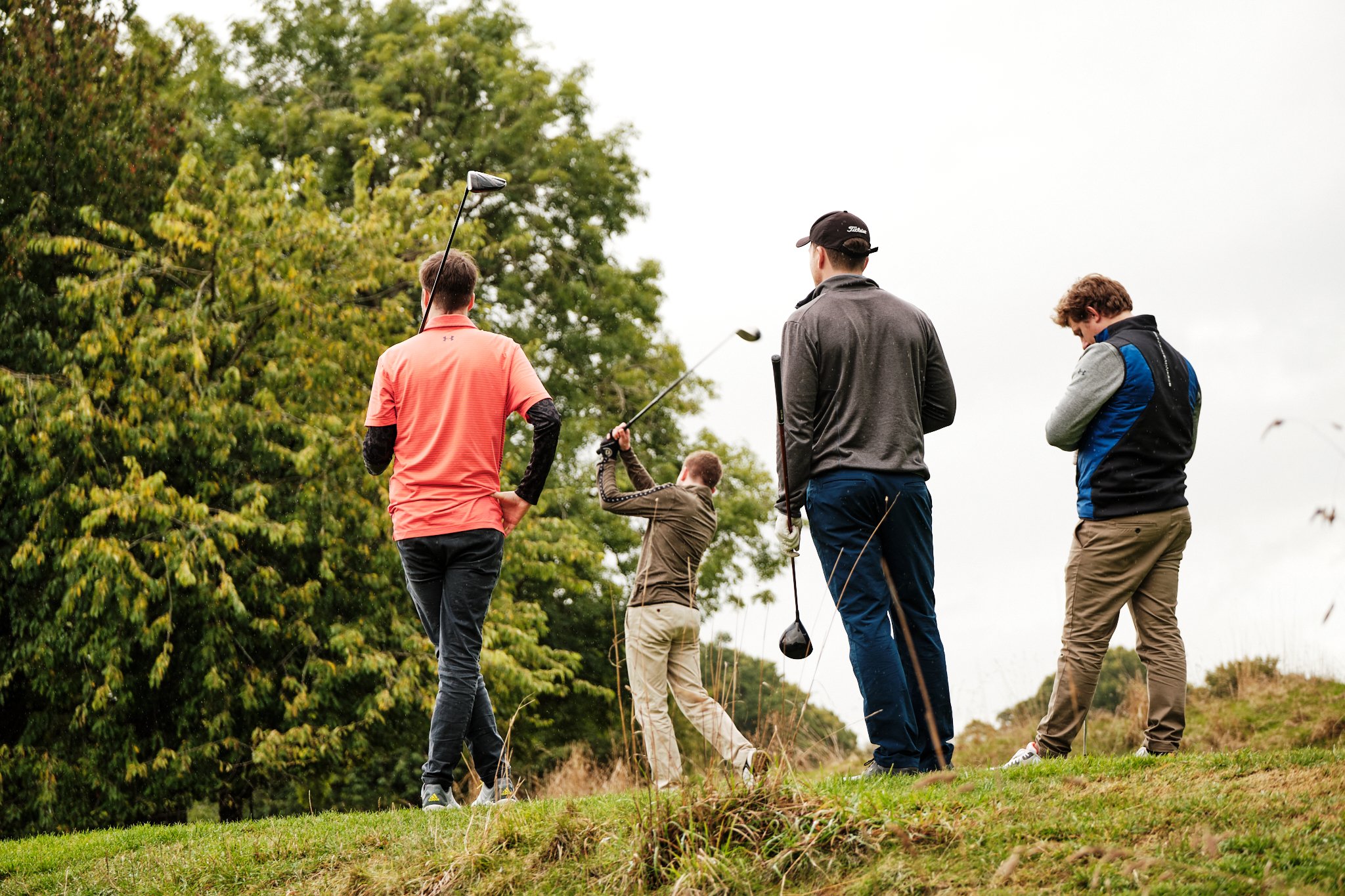 Four men standing on a grassy area with trees in the background, one swinging a golf club.