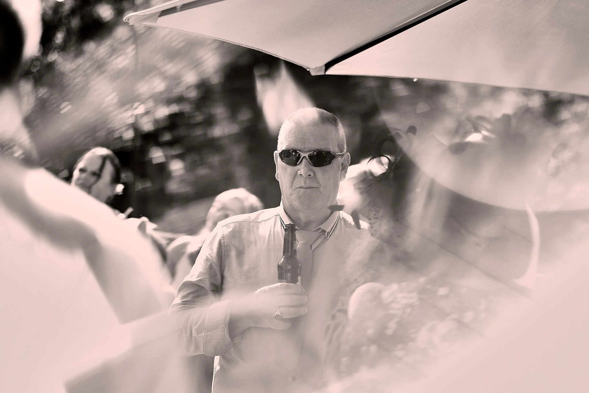 A man with sunglasses holding a beer bottle and taking a selfie, seen through a reflective surface, with a blurred background.