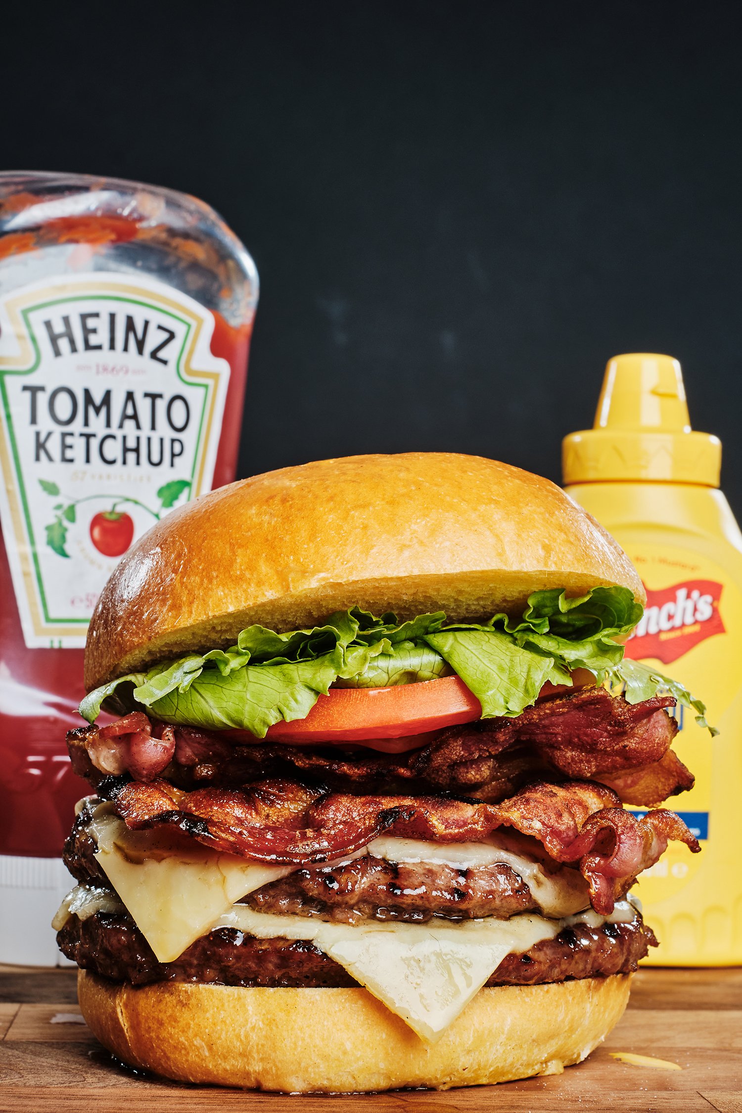 Close-up of a large burger with bacon, cheese, lettuce, tomato, and a bun, with ketchup and mustard bottles and Heinz ketchup bottle in the background.