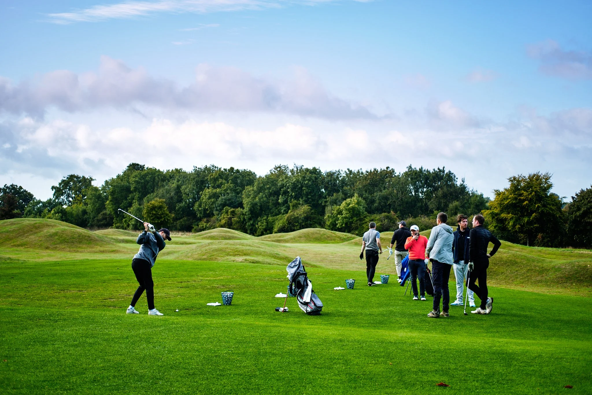 A group of people practicing golf at a golf course, with one woman swinging a golf club in the foreground and others standing and talking in the background.