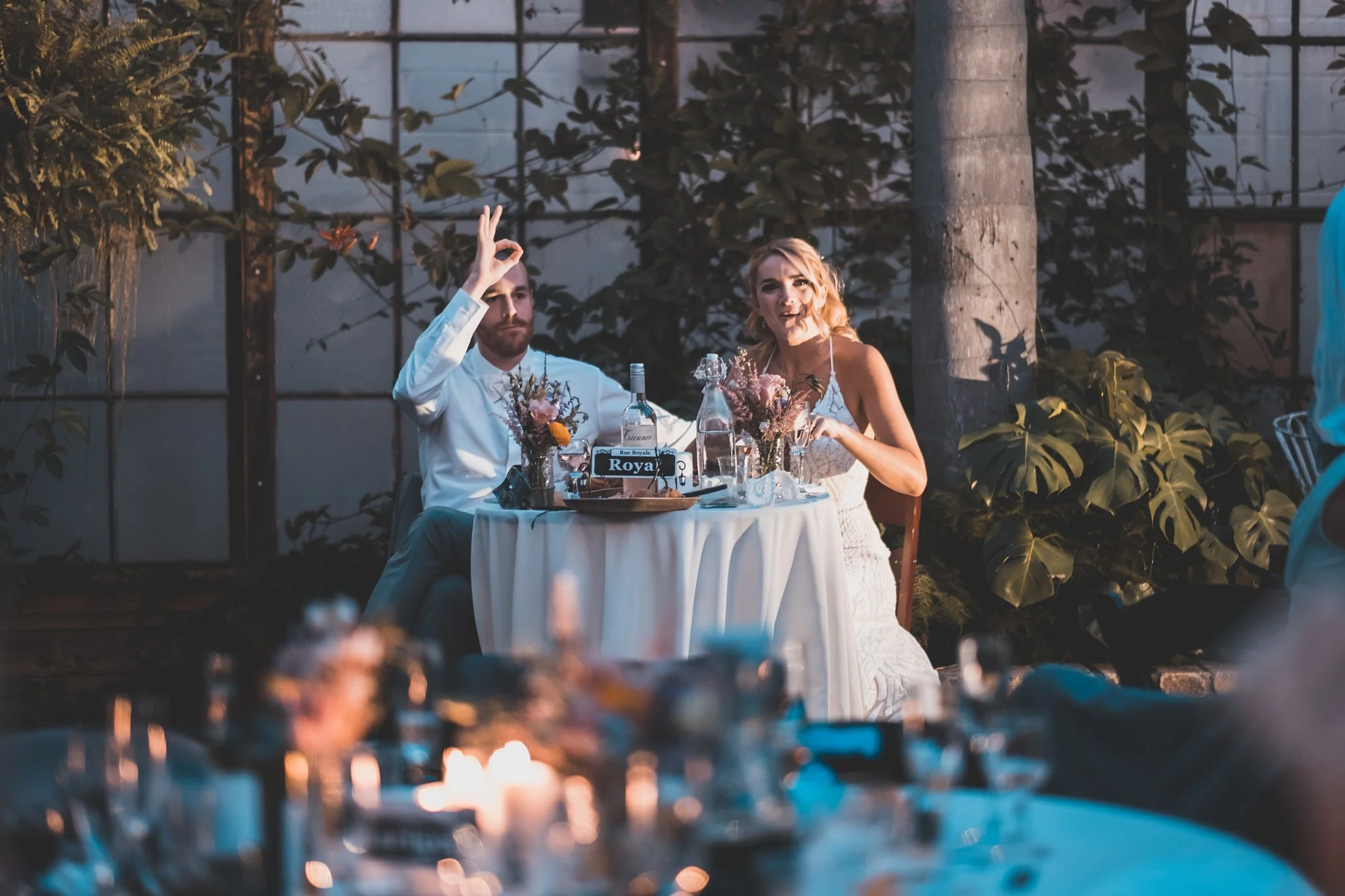 A couple celebrating at a wedding reception, sitting at a table with floral centerpieces and bottles of alcohol, outdoors surrounded by greenery and a tree, during the evening.