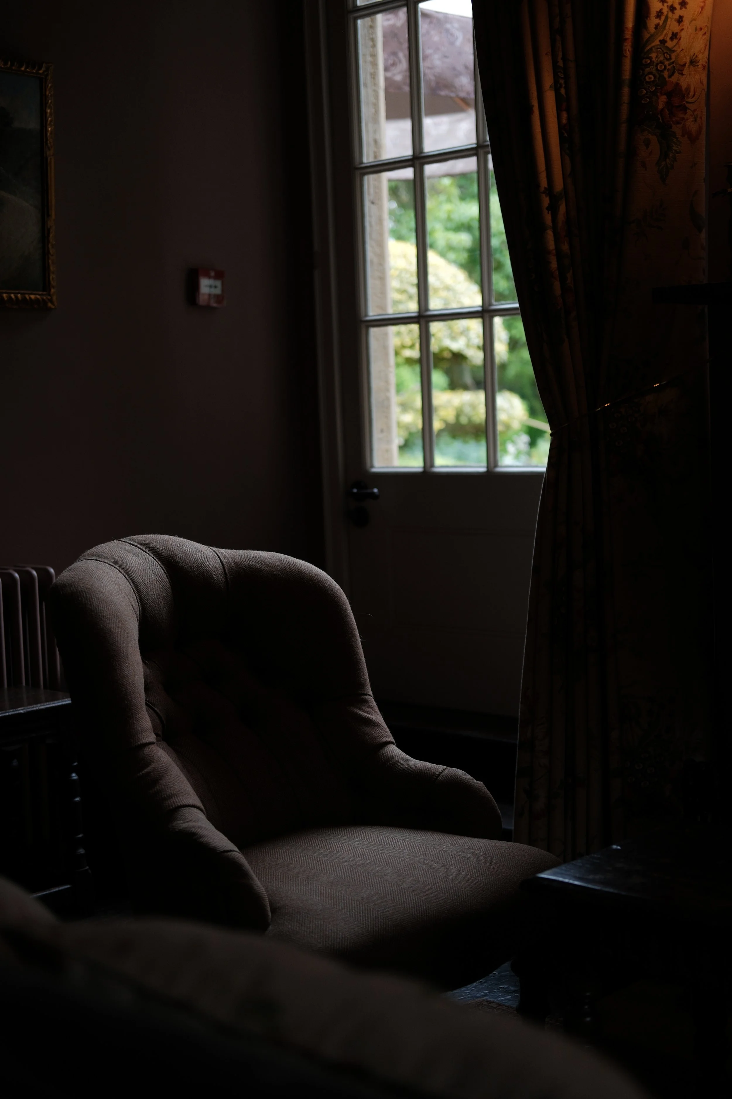 Dimly lit room with a cushioned armchair near a window showing green trees outside, partially covered by curtains.