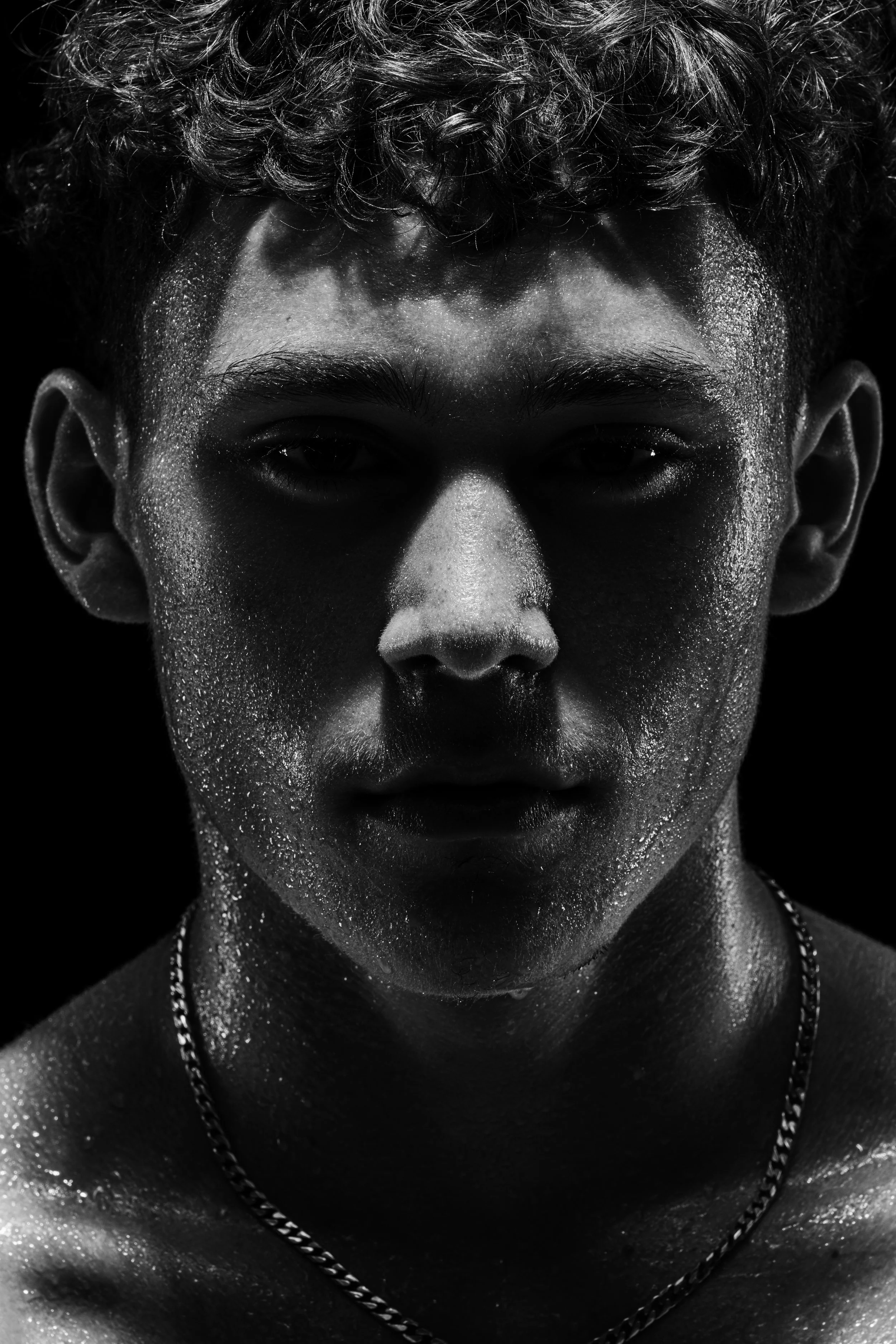 Close-up black and white portrait of a young man with curly hair and shiny skin, wearing a chain necklace, looking directly at the camera.