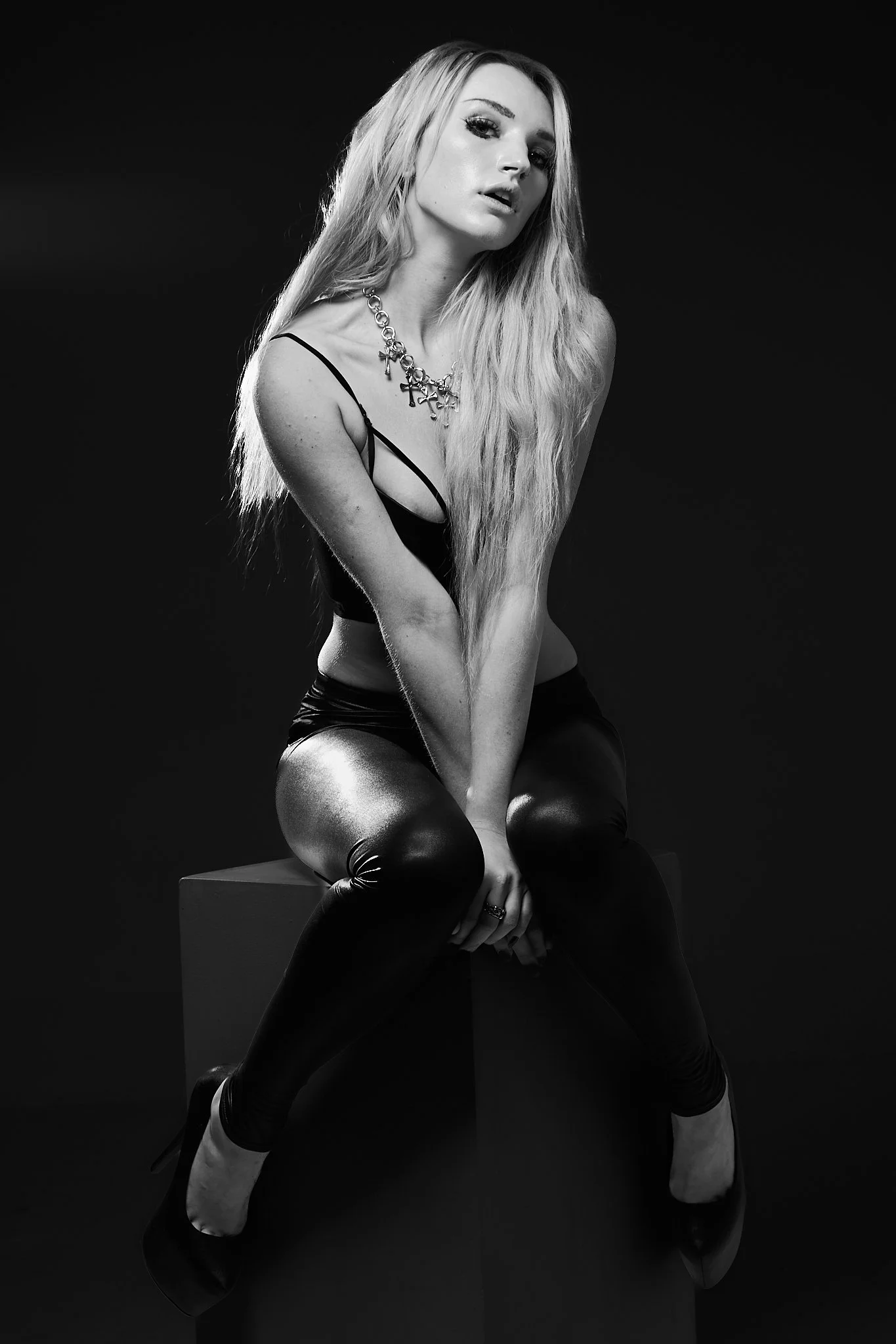 Black and white photo of a woman sitting on a block, wearing a black camisole, shiny black pants, high heels, and a chunky necklace, with long wavy blonde hair and a serious expression.