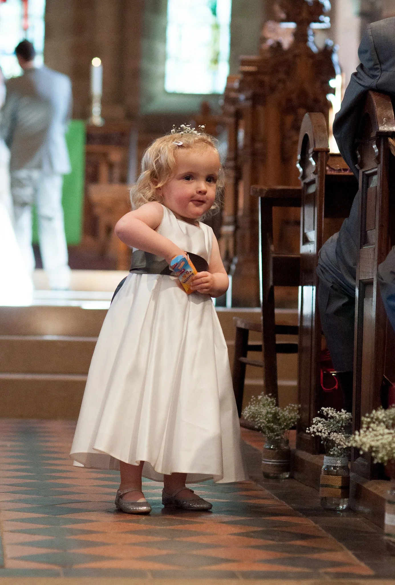 Young girl with curly blonde hair wearing a white dress and silver shoes, standing inside a church near the altar, holding a small object in her hand, with flowers on the floor nearby.