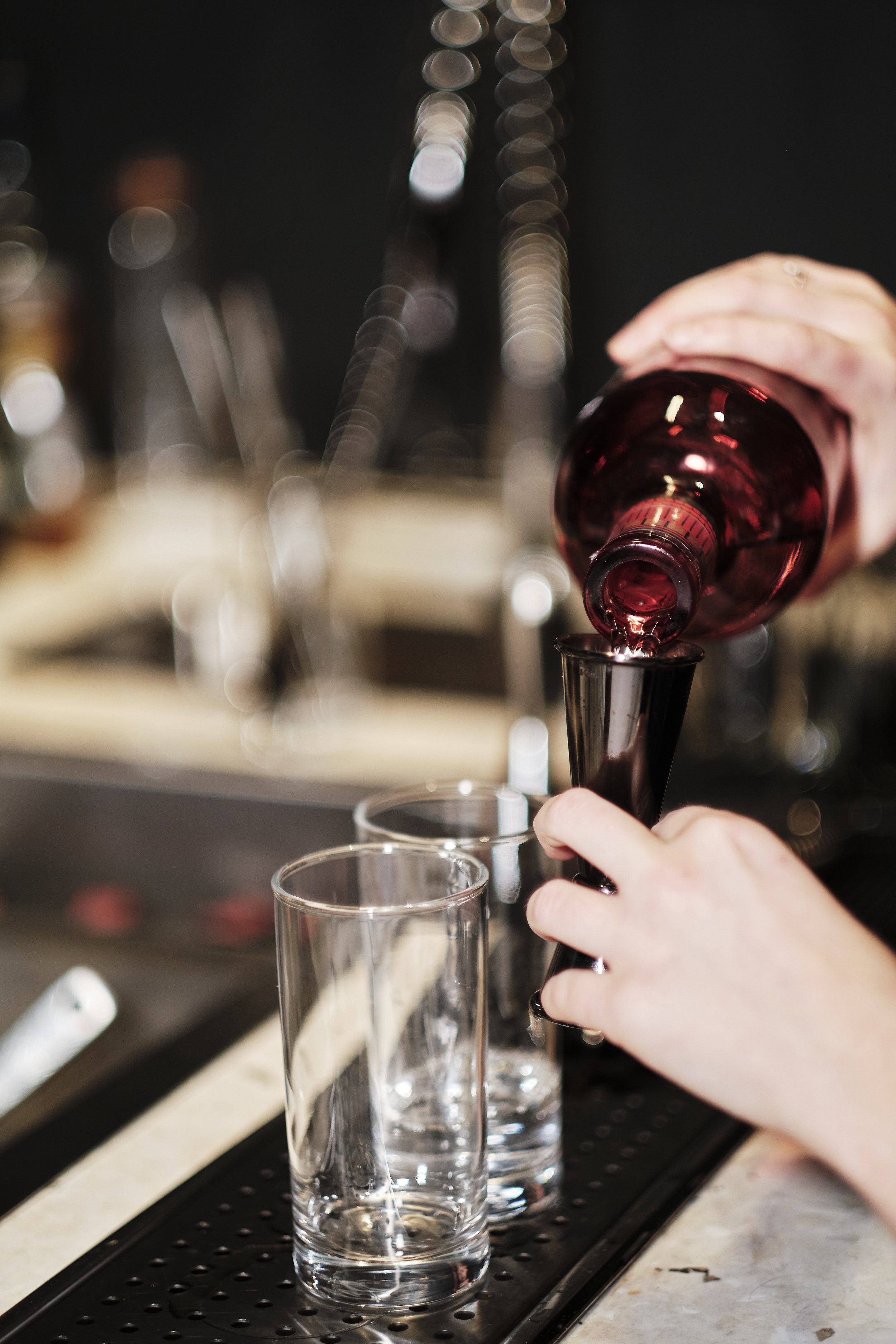 A bartender pouring red wine from a bottle into a small glass, with other glasses on a bar counter.