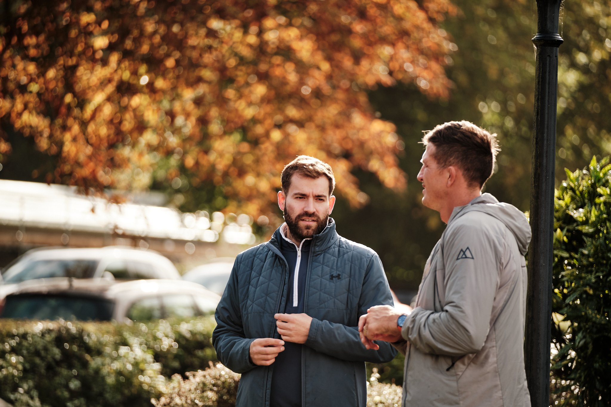 Two men talking outdoors on a sunny day with autumn foliage in the background.