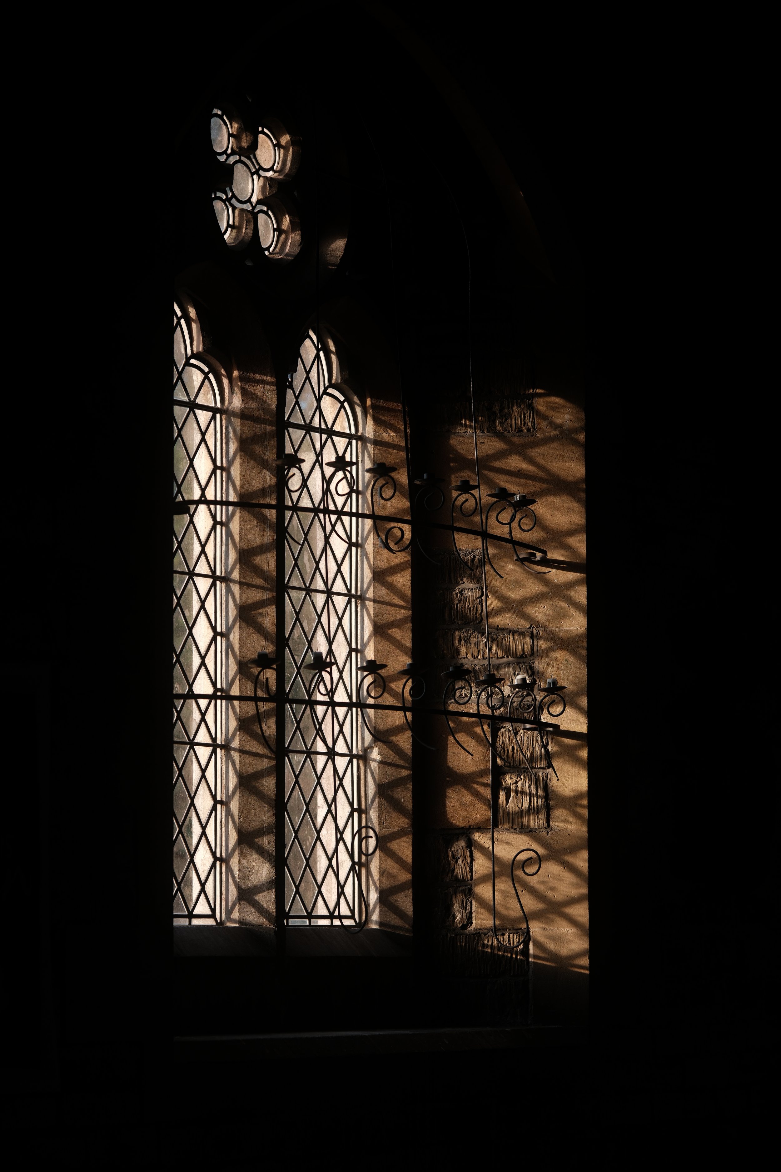 Sunlight streams through a decorative, wrought-iron window with diamond-shaped panes, casting intricate shadows inside a stone building.