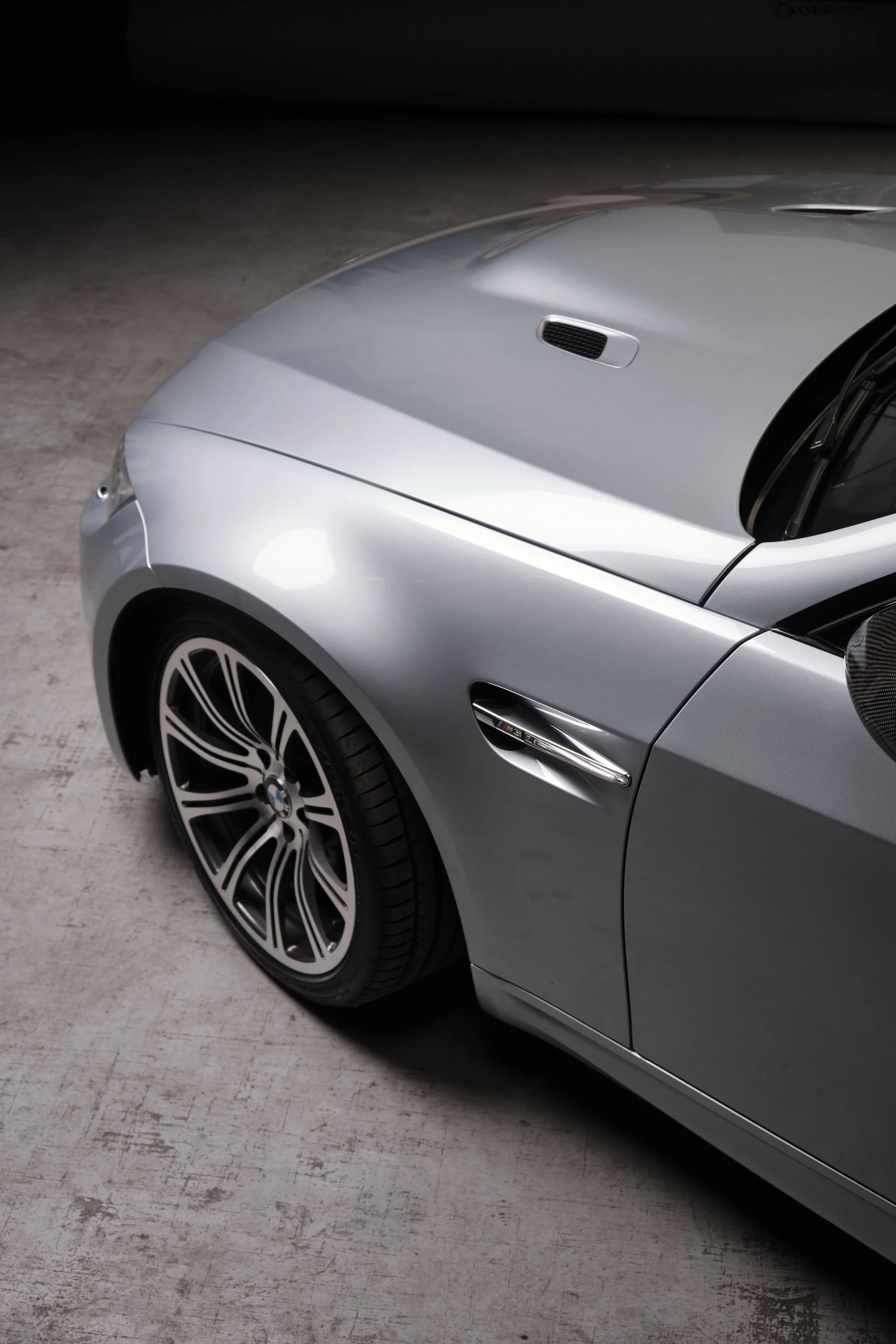 Close-up of a silver BMW sports car's front left side, showing the wheel, headlight, and side vent with an M badge.