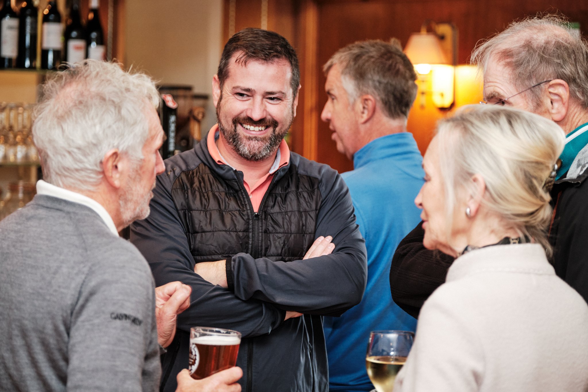 A group of six adults socializing at a bar or pub, with drinks in hand, engaging in conversation and smiling.