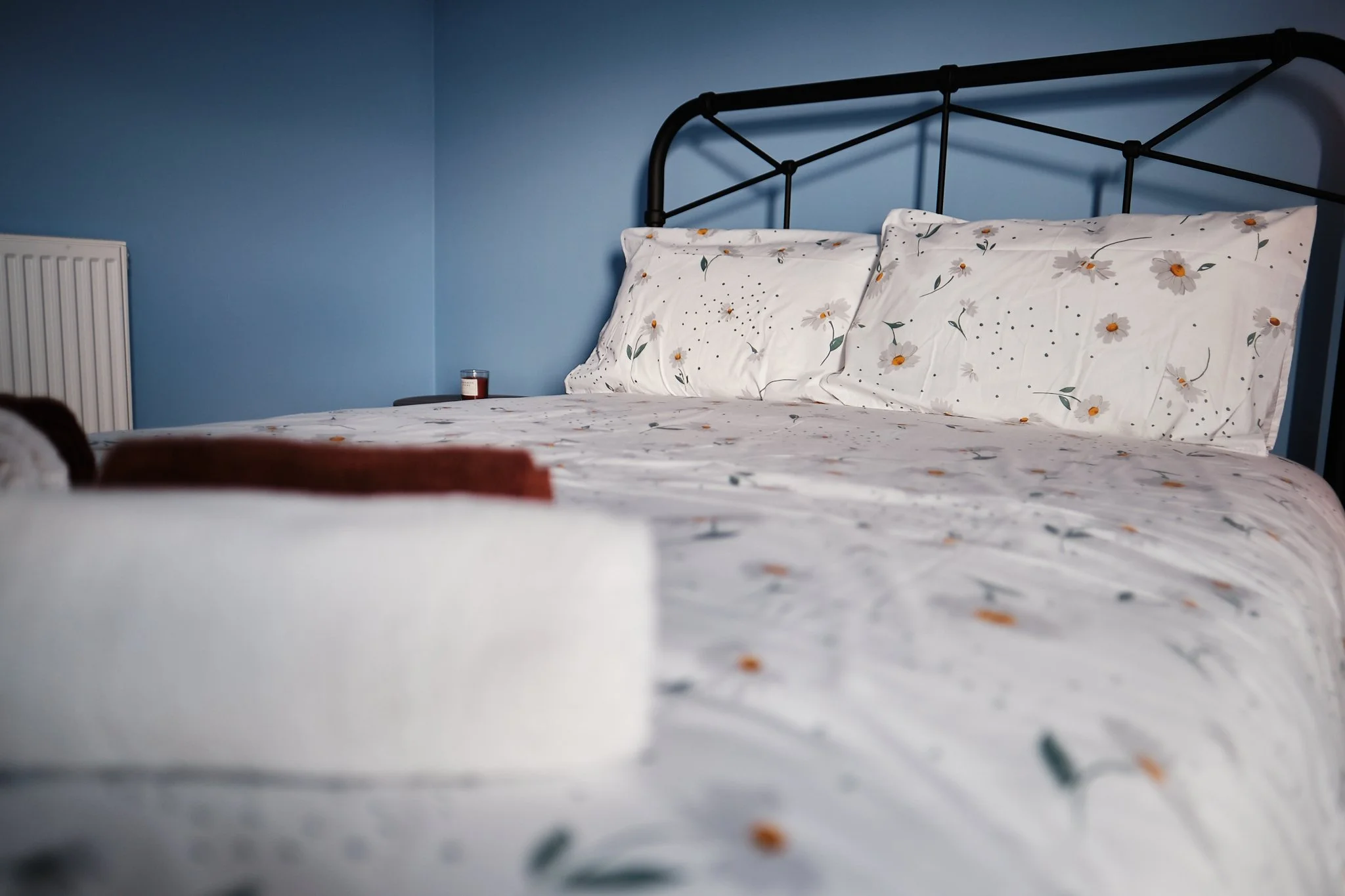 A neatly made bed with floral sheets and pillows, set against a blue wall with a radiator on the side.