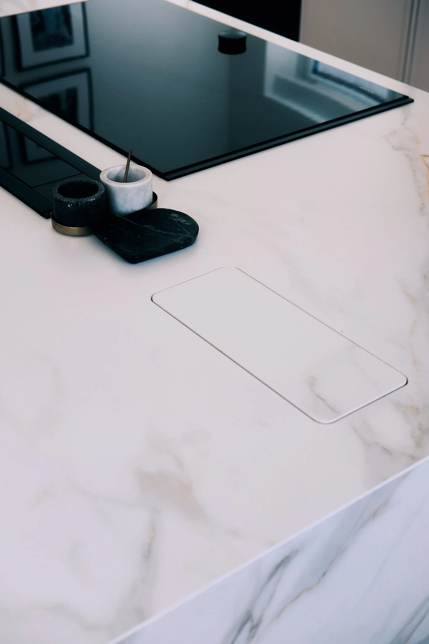A modern kitchen countertop with a built-in induction cooktop, a white marble surface, and a small tray containing black, gray, and white containers.