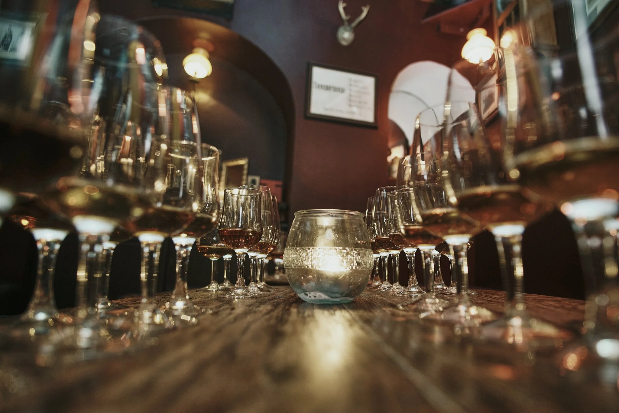 Wine glasses arranged on a rustic wooden table in a dimly lit restaurant, with a candle in a glass holder at the center and a clock, deer head, and framed pictures on the dark-colored walls.