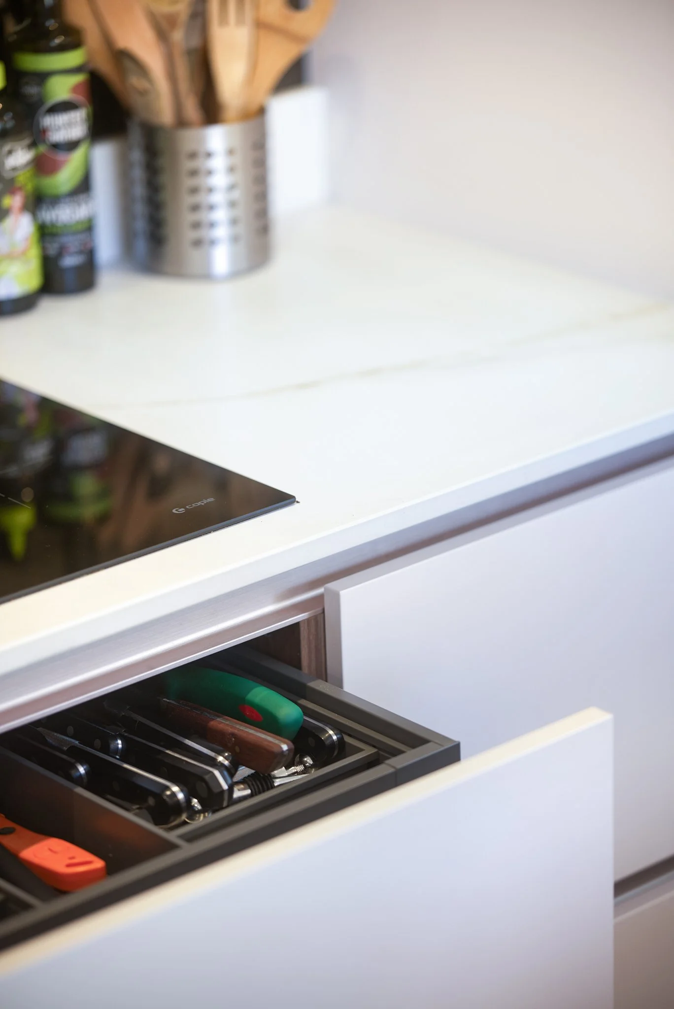 Kitchen countertop with an induction stove and an open drawer containing knives and kitchen tools, with a container holding wooden utensils and spice bottles in the background.